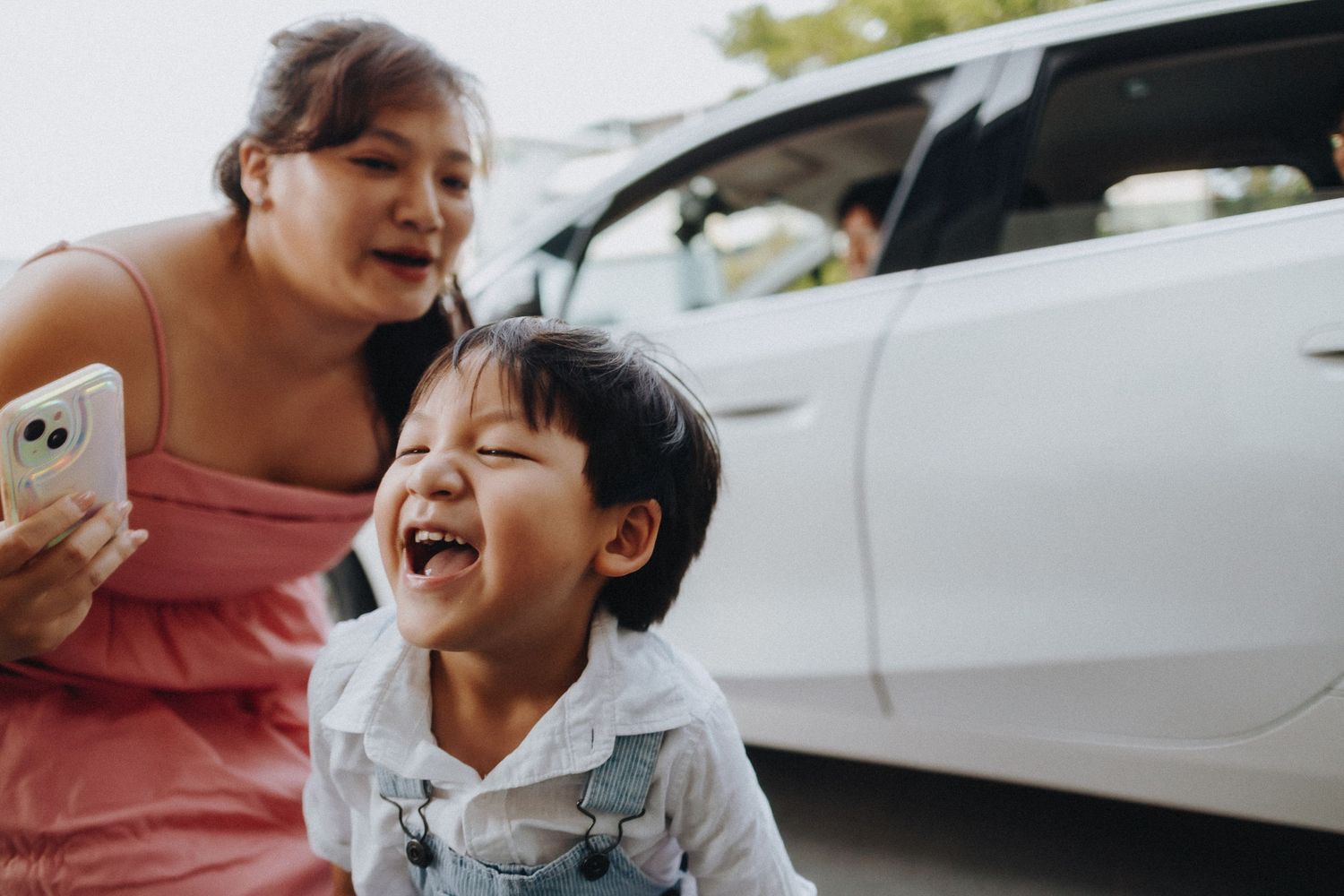 A young child laughs joyfully while wearing a denim jacket, standing next to a car outdoors.
