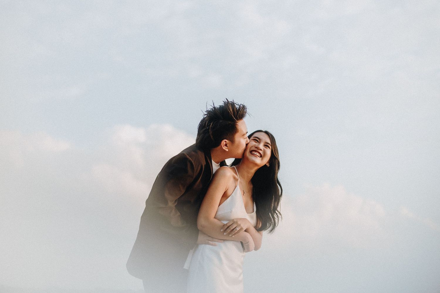 A joyful wedding couple laughs together against a cloudy sky in an intimate moment.