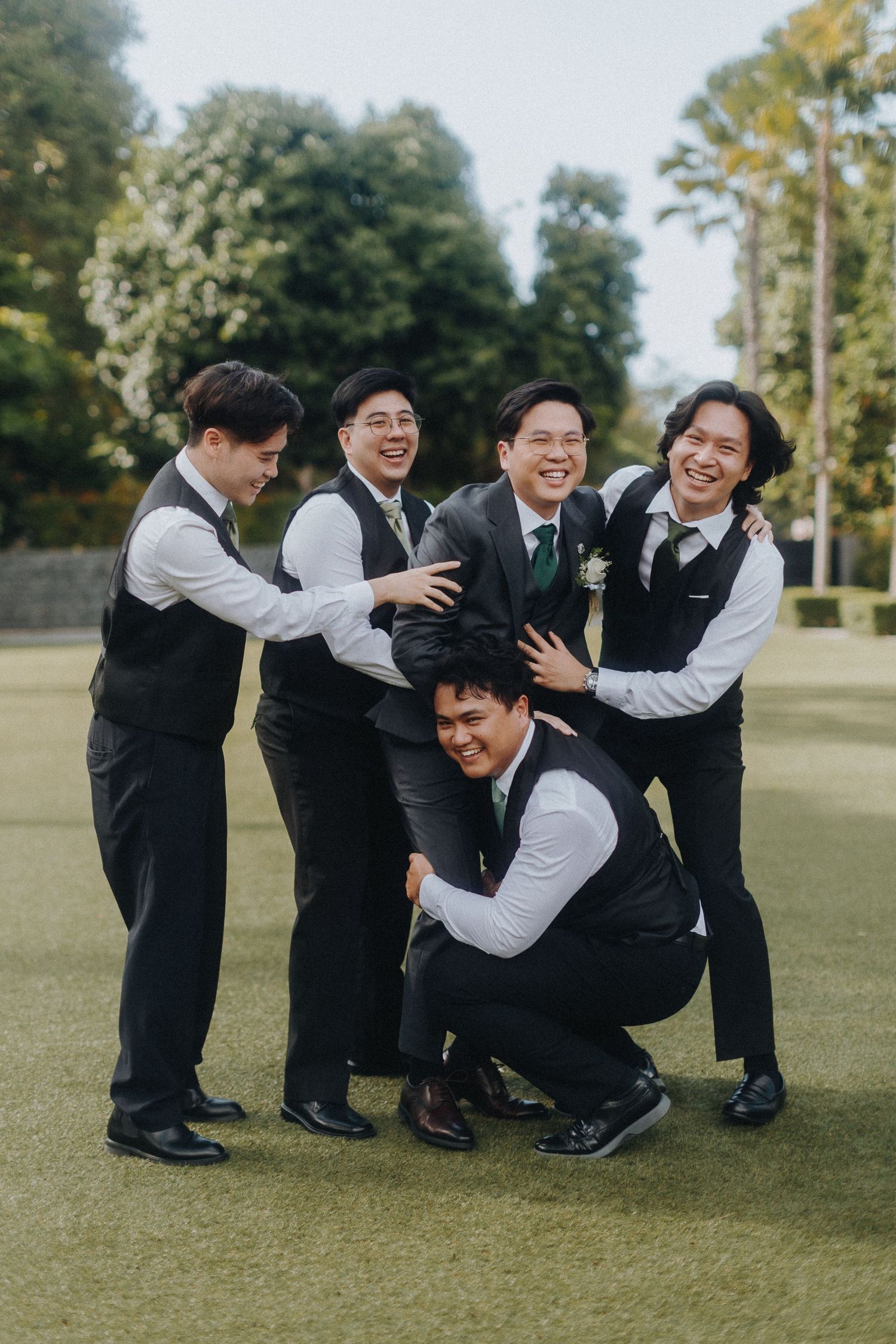 Groomsmen in matching vests sharing a playful moment on a green lawn during wedding festivities.