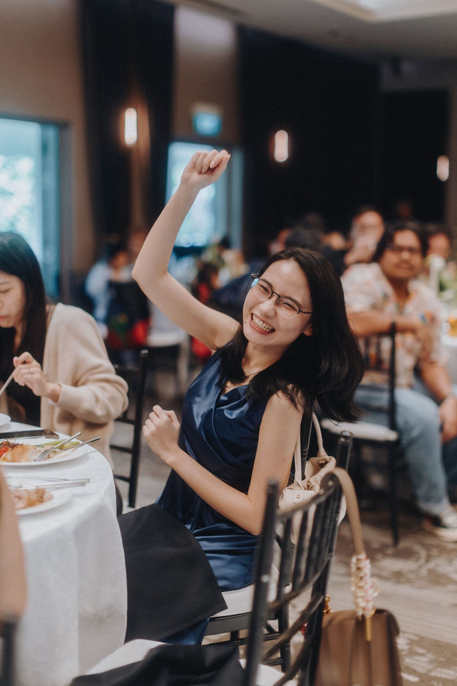 A woman in a navy blue dress enthusiastically raises her arm in celebration during a restaurant gathering.