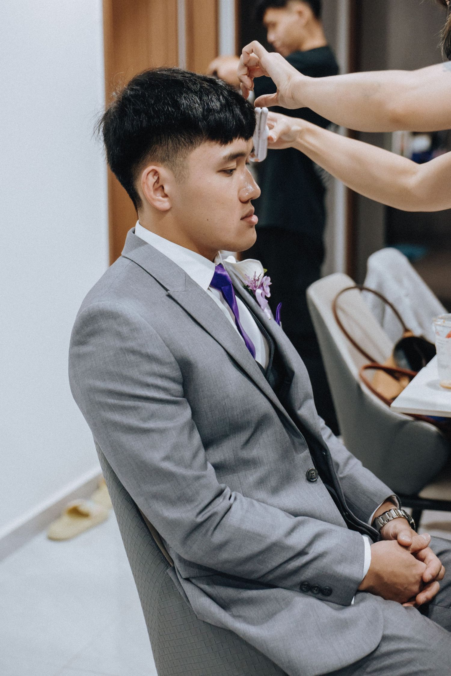 A person in a grey suit with a purple tie getting their hair styled while seated in a chair.