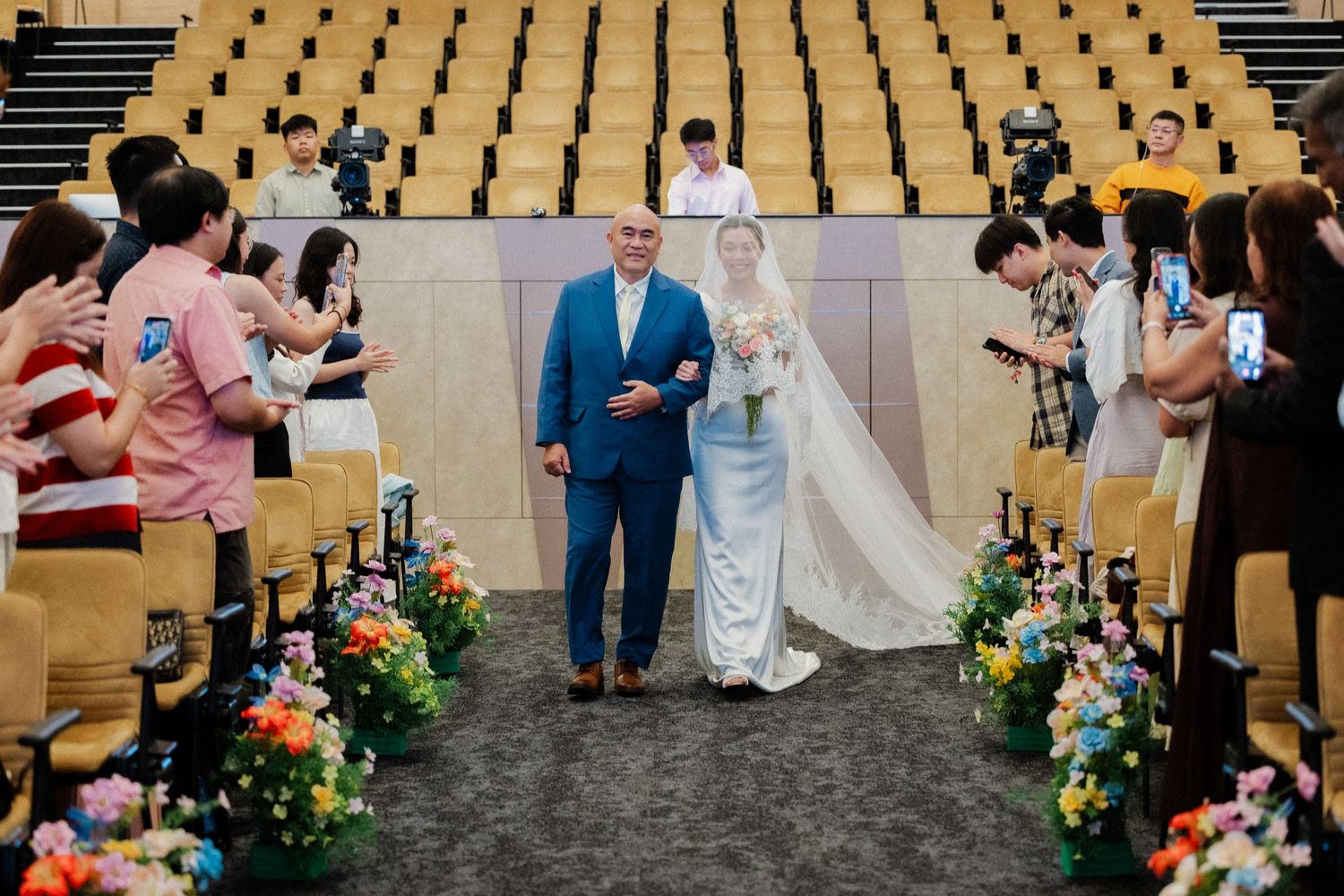 Bride in full veil walks down auditorium aisle escorted by father, flanked by colorful floral arrangements.