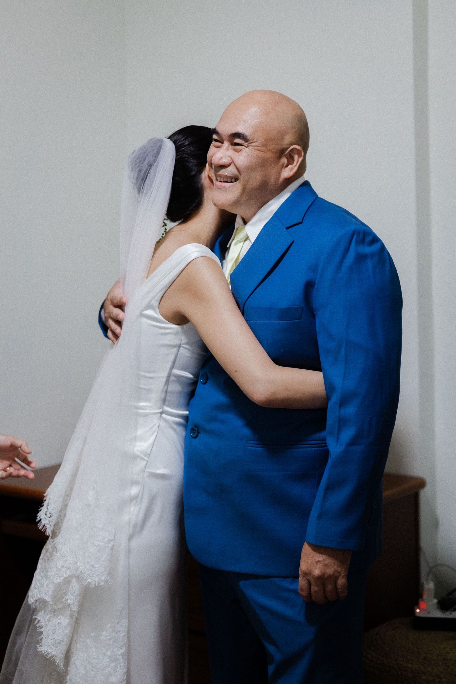 Bride in white gown hugging a smiling bald man in a blue suit during a wedding celebration.