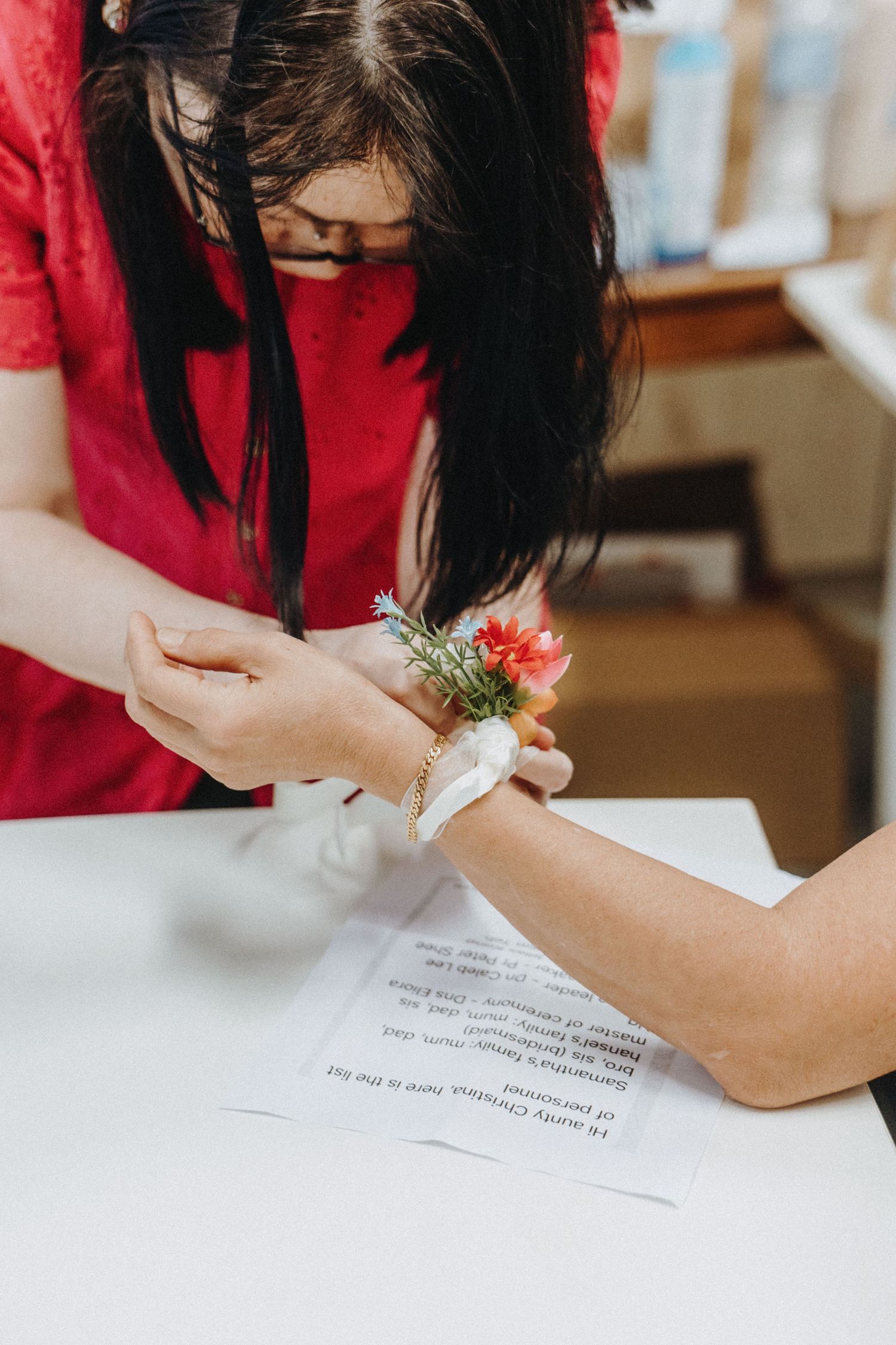 Woman in red dress attaching a floral wrist corsage with red flowers onto another persons wrist at a wedding.