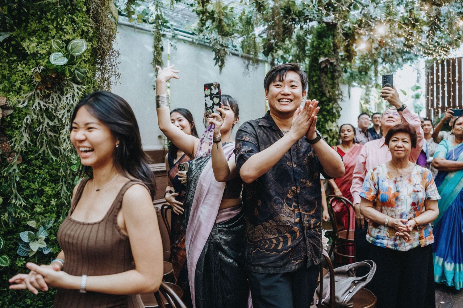 Happy guests in traditional attire cheering and clapping at a lush outdoor wedding celebration.