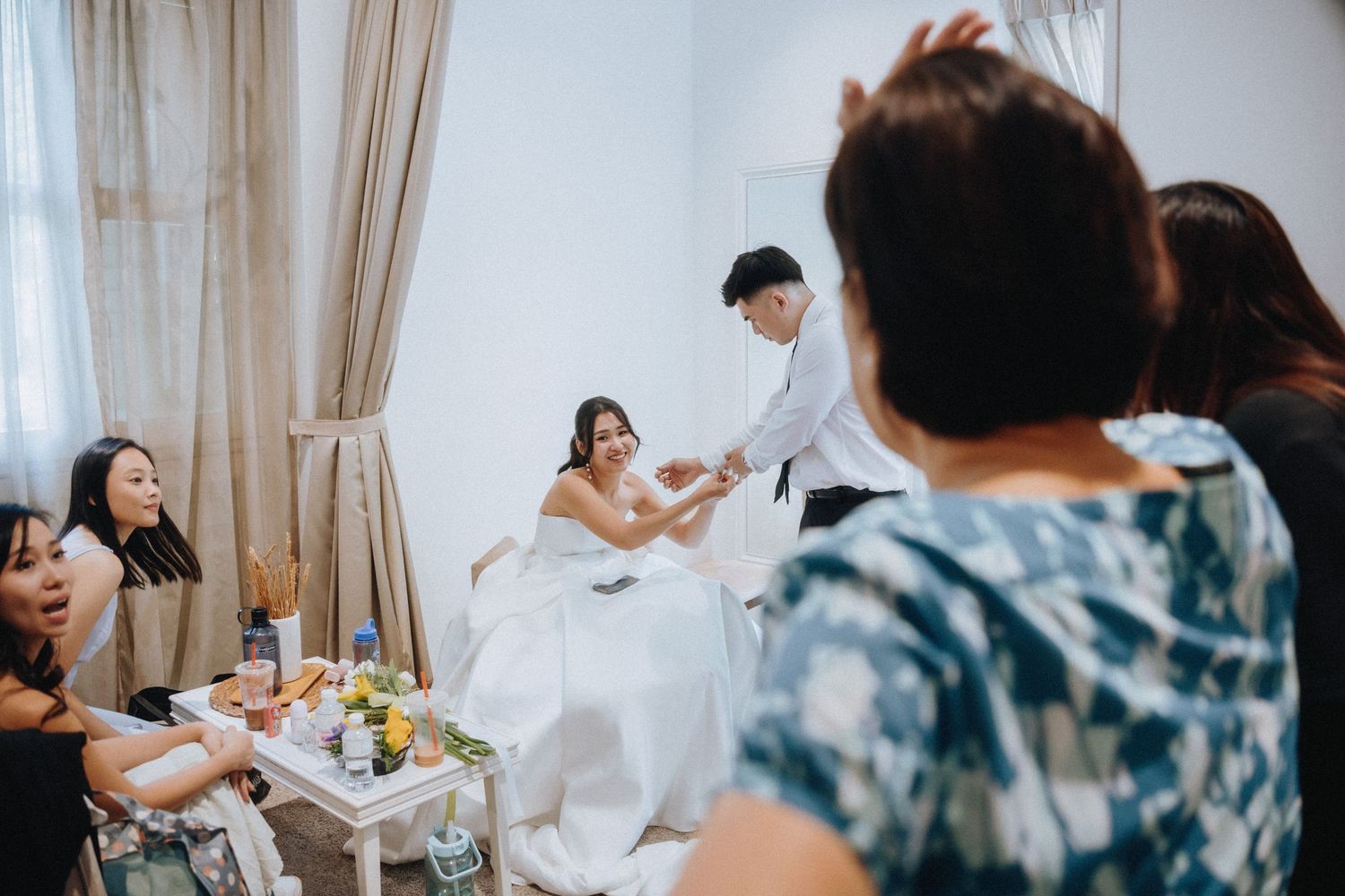 Bride in white gown surrounded by family and friends during wedding preparations in bright room.