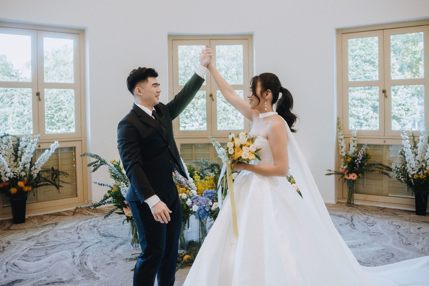 Bride and groom high-five joyfully during wedding ceremony in bright white venue with flowers.