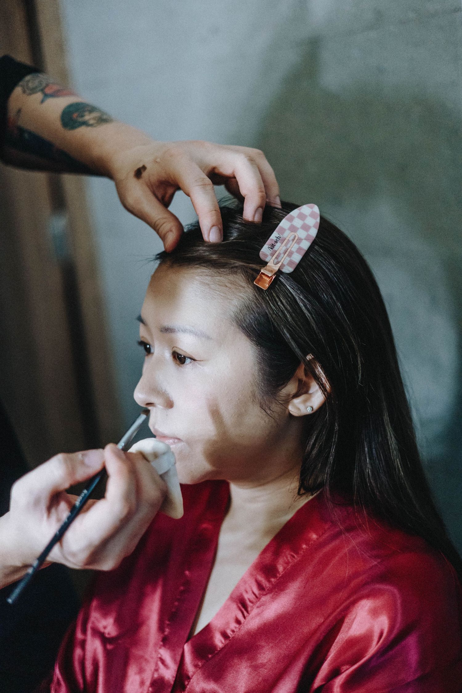 A makeup artist applies lipstick and styles hair with a clip on an Asian woman in a red satin robe.