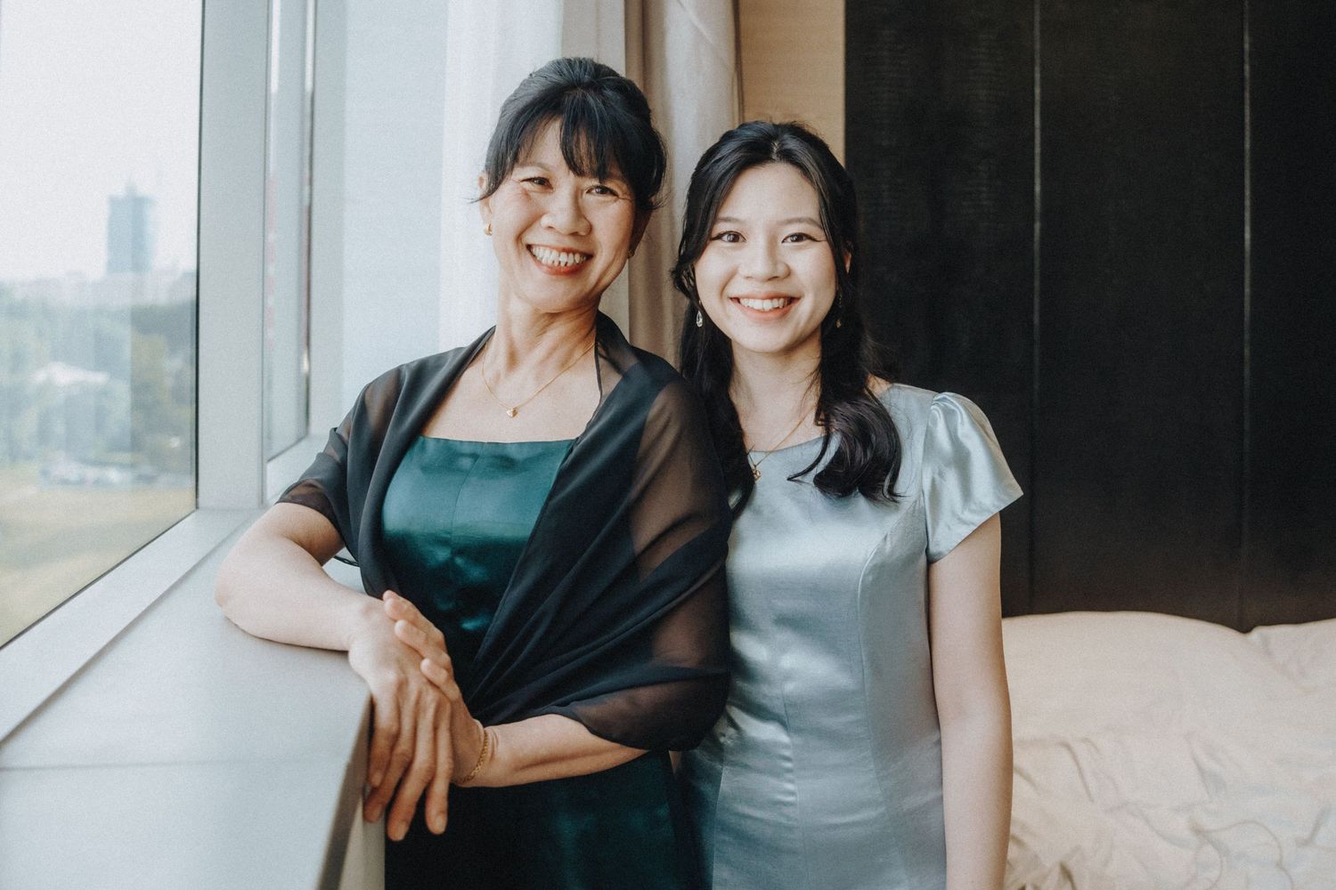 Two smiling women in elegant dresses pose together by a bright window in a modern interior setting.