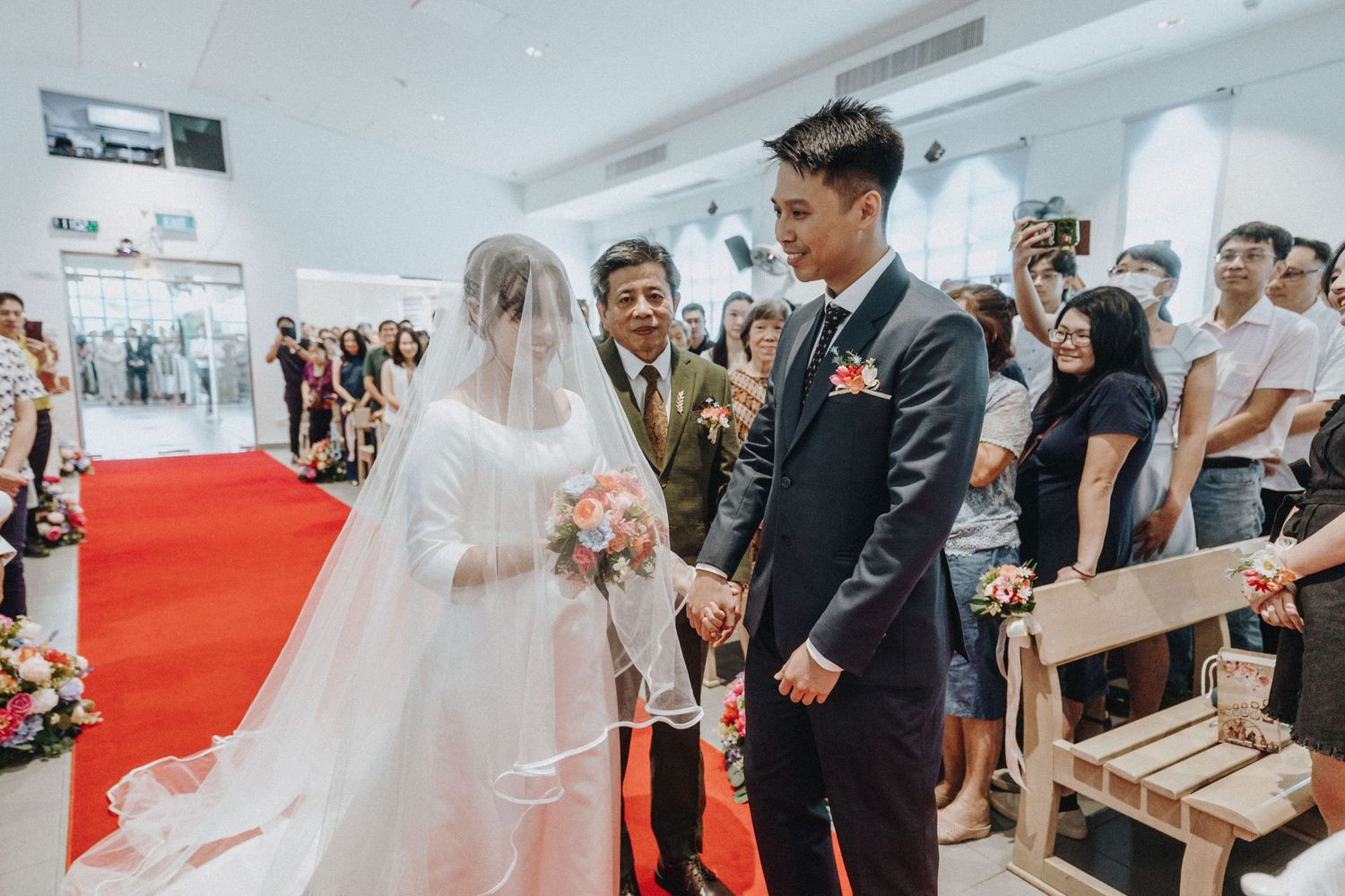 A bride in a white veil and gown holds hands with a groom in a black suit at a church ceremony.