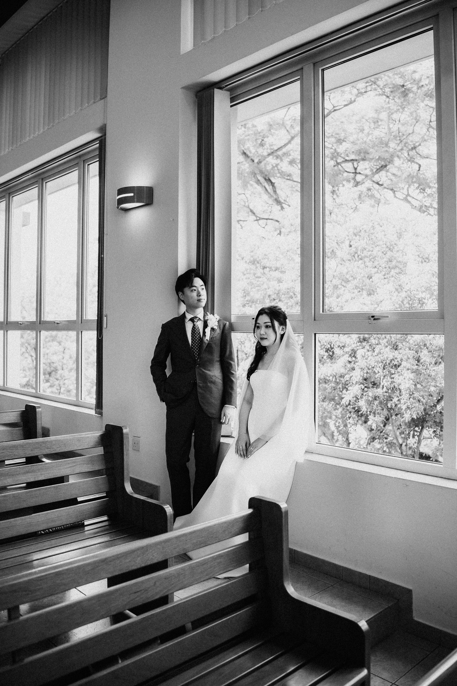 Black and white photo of bride and groom posing by large windows in a church stairwell on their wedding day.