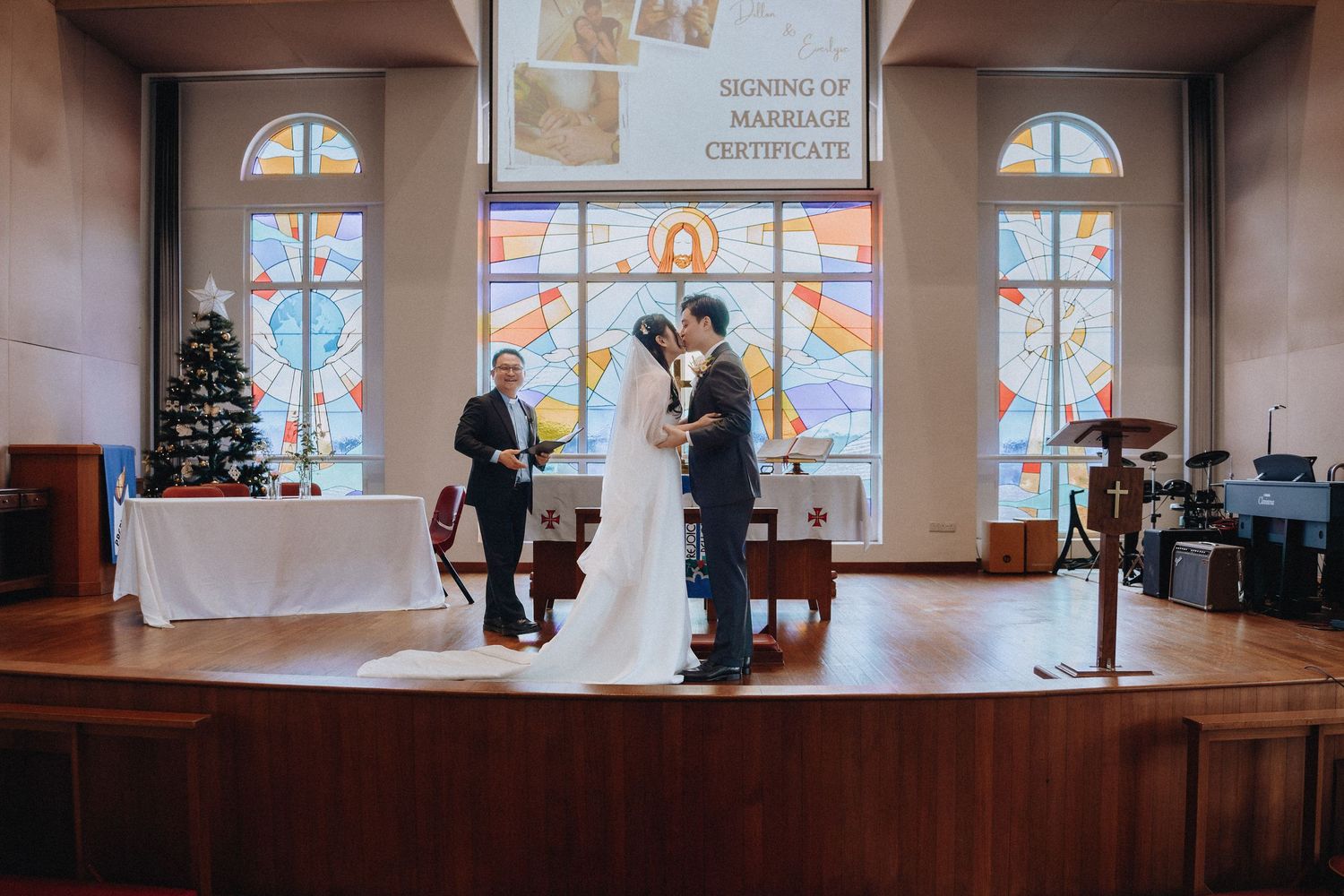 Couple sharing a kiss at church altar during wedding ceremony with stained glass windows and marriage certificate sign.