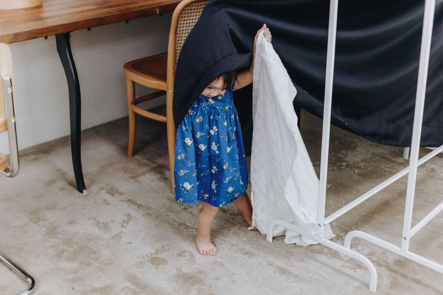 Small child in blue floral dress peeks out from behind white curtain in minimalist room setting.