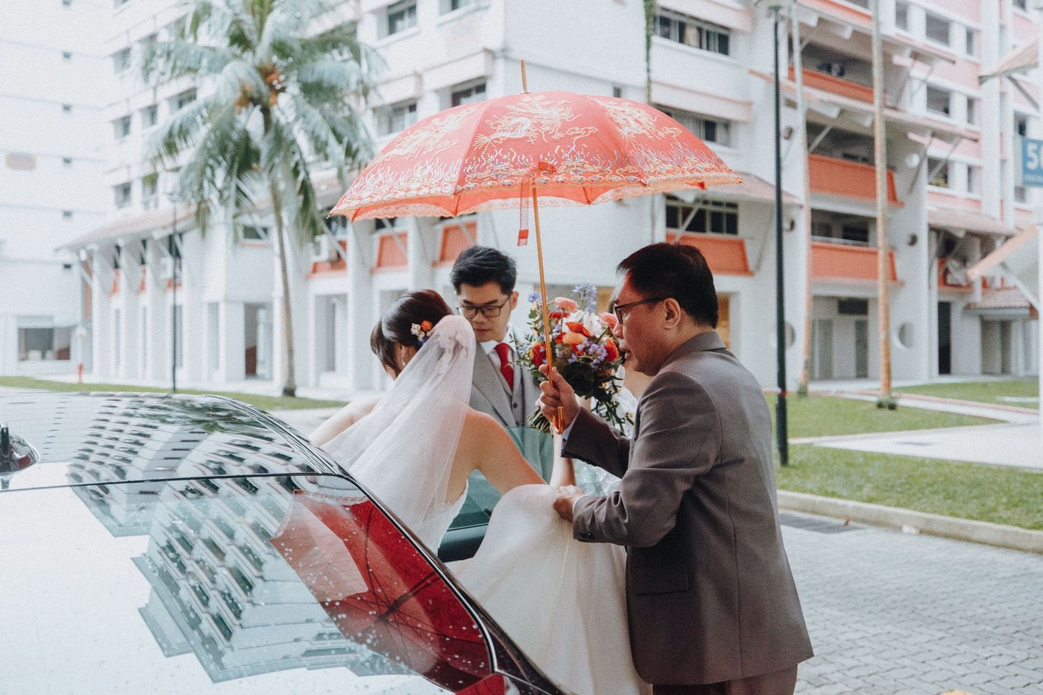 Wedding couple getting into car while helpers hold red umbrella on rainy day.
