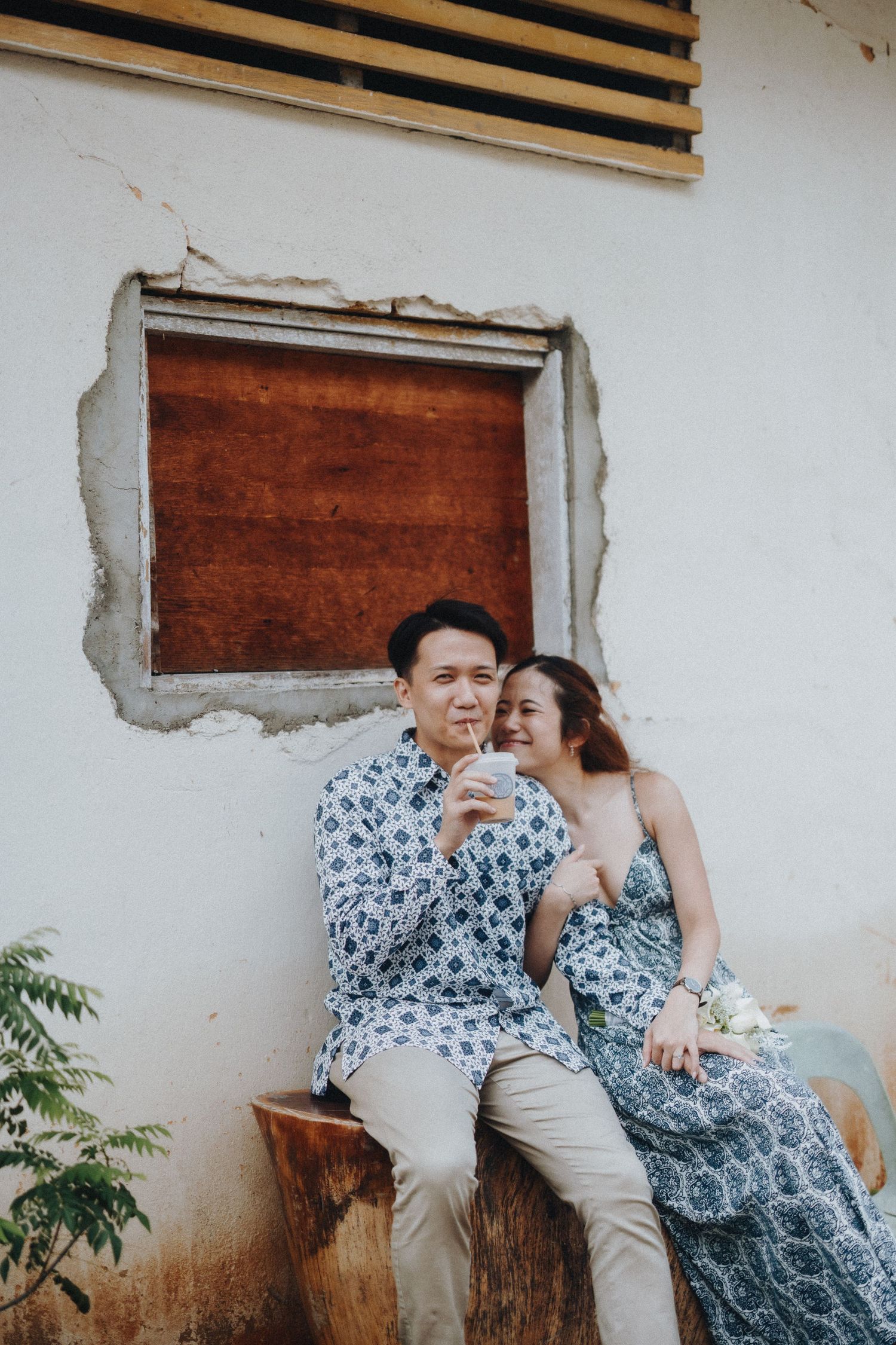 A couple in matching blue floral outfits sits together against a rustic white wall in a romantic outdoor setting.