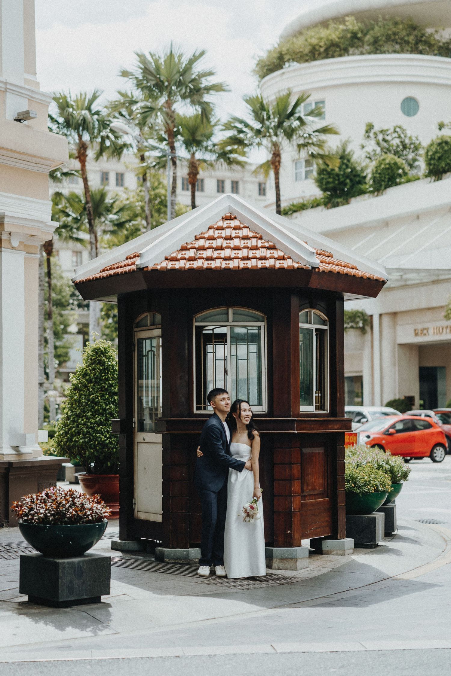 A couple poses by a vintage wooden kiosk with terracotta tiled roof outside a white building with palm trees.