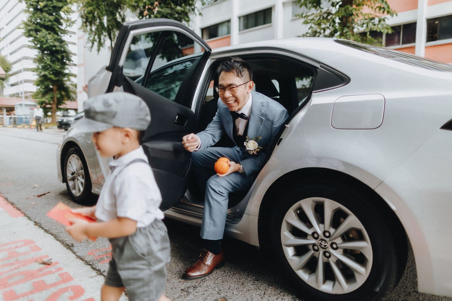 A sweet interaction between a child in white and an adult by a silver car on a city street.