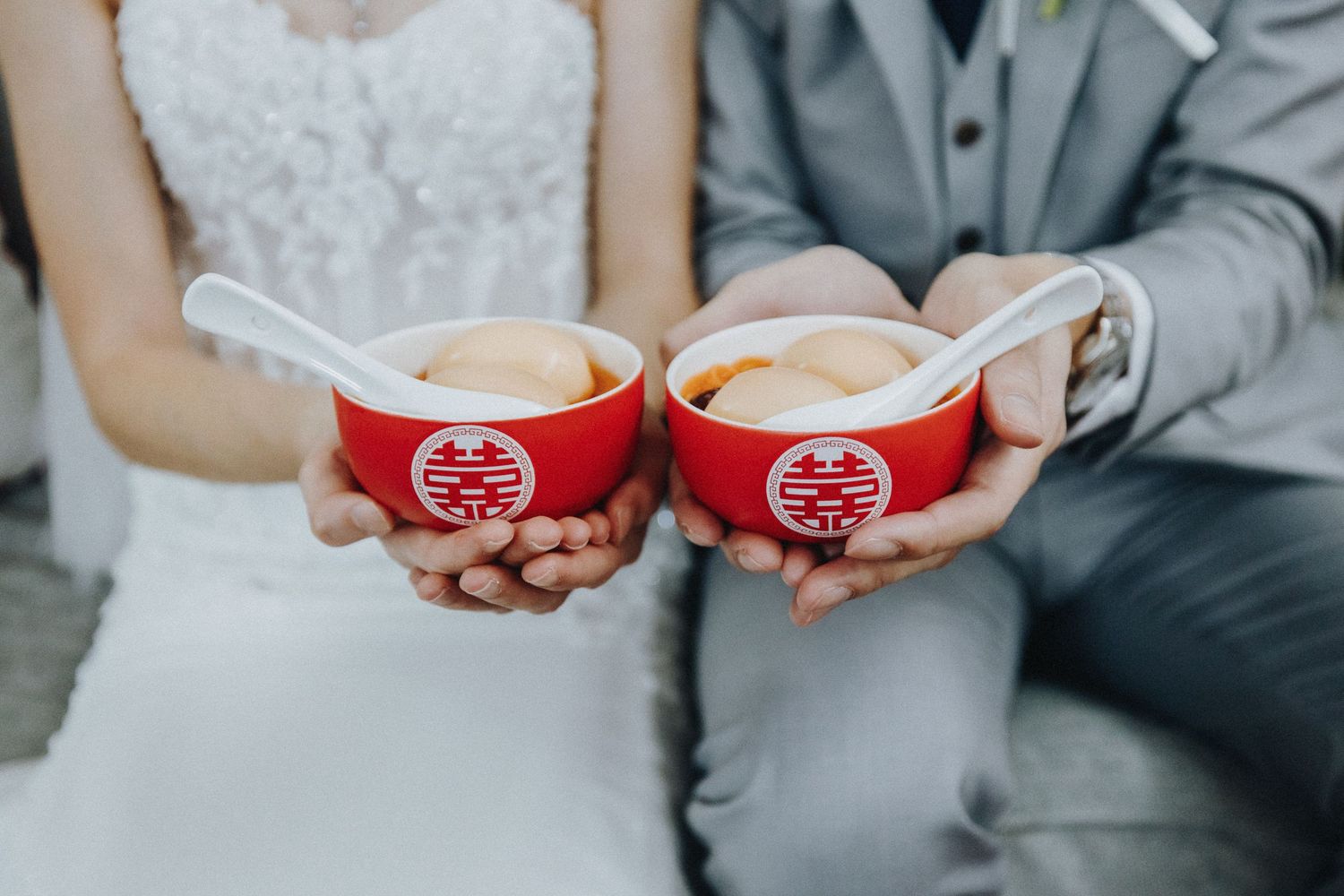 Wedding couple holds traditional red Chinese tea bowls with double happiness symbols for tea ceremony ritual.