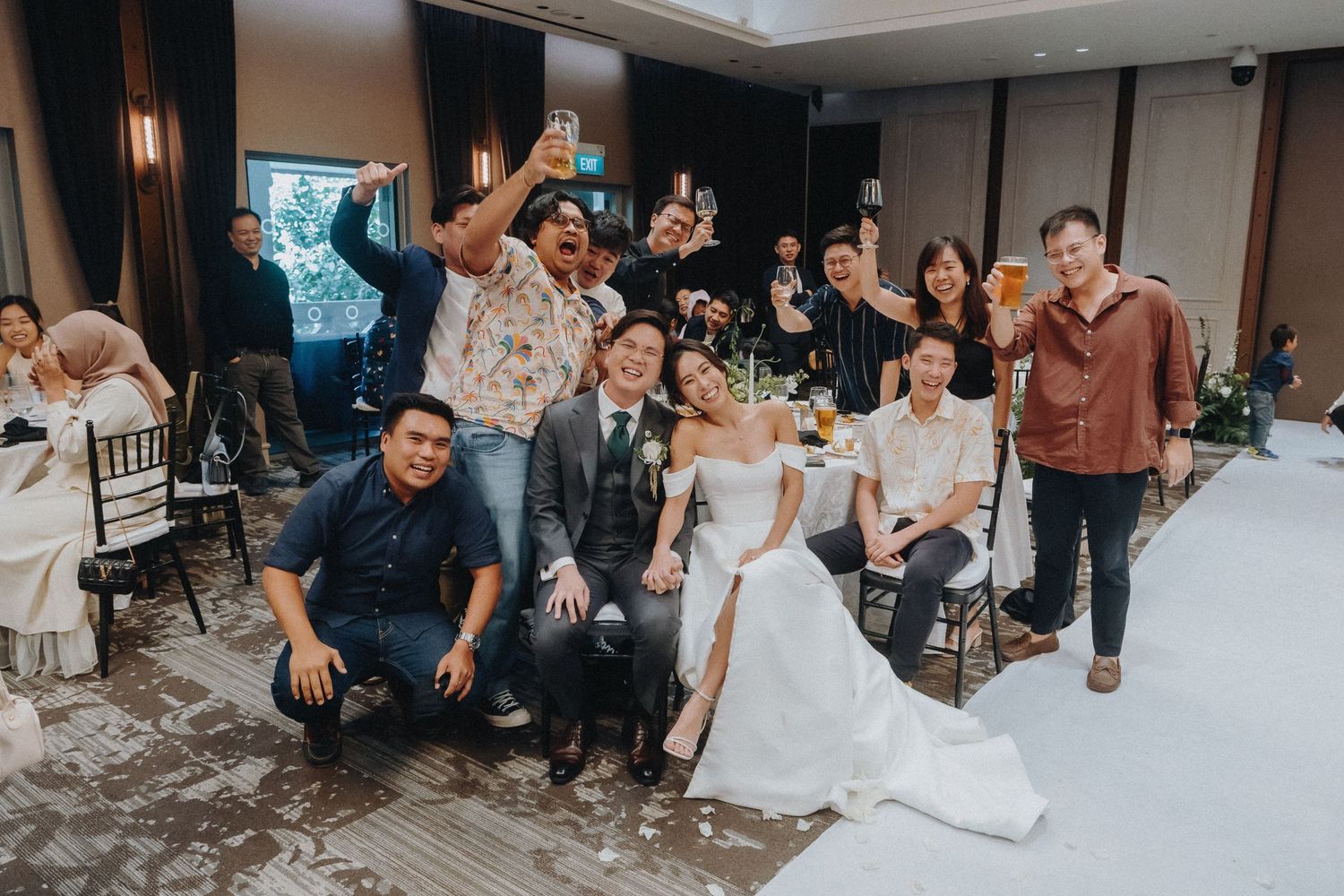 A joyful group photo of wedding guests and the newlyweds posing together in a reception hall with raised arms.