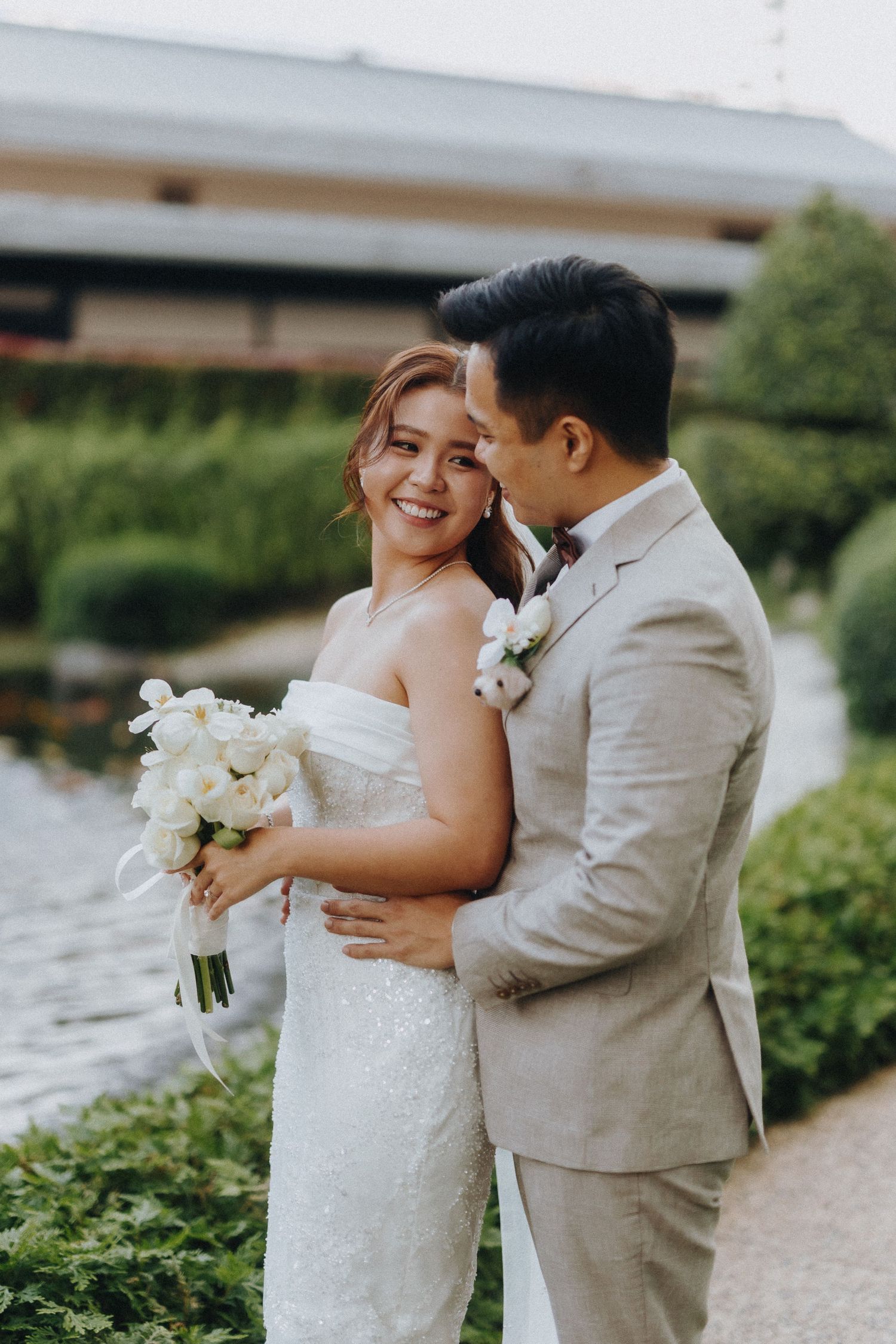 A couple shares an intimate moment by the water, with the bride holding a white floral bouquet.
