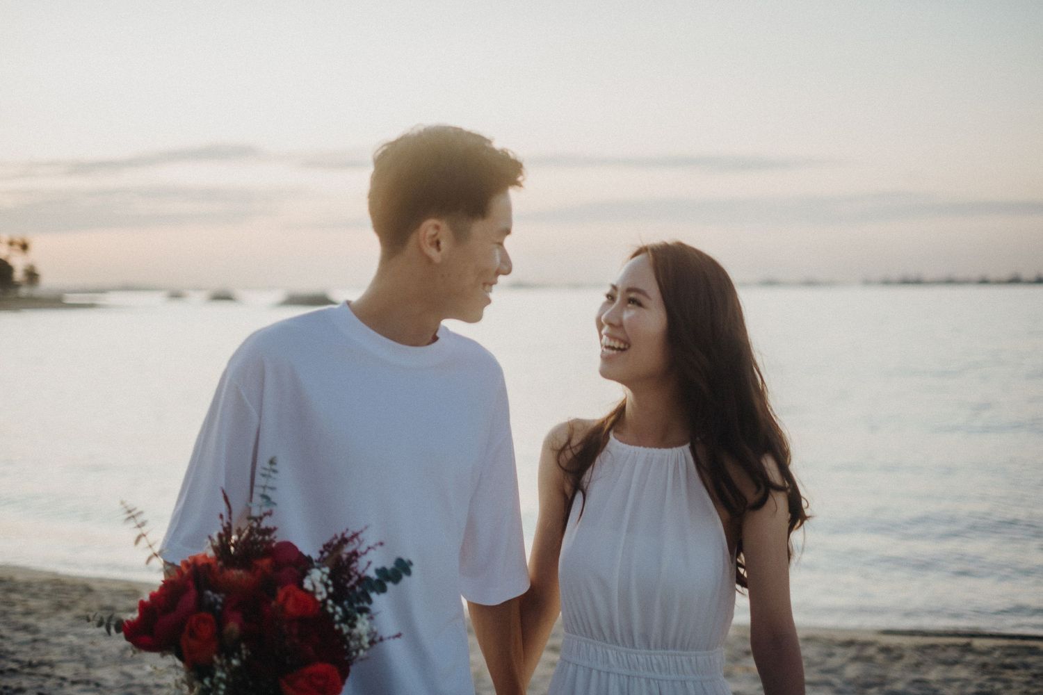 Couple shares tender moments during sunset beach photoshoot with red floral bouquet in Hawaii.