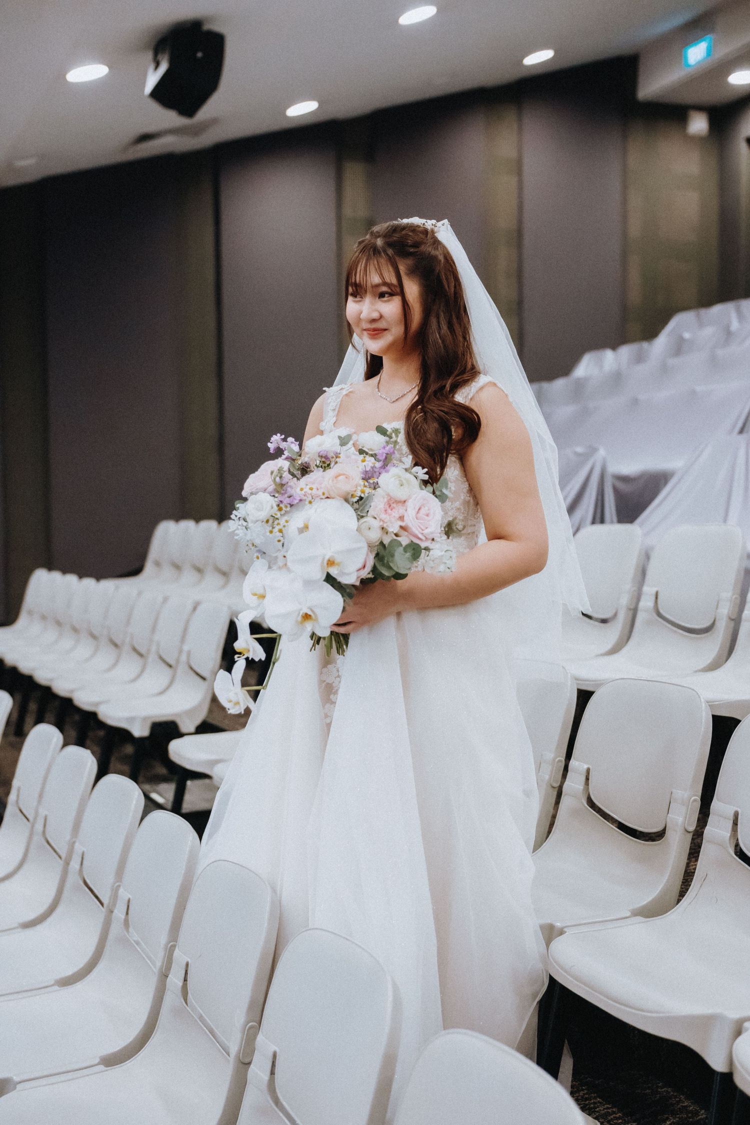 Bride in white dress holding a pink and white floral bouquet stands among rows of white chairs in an indoor venue.
