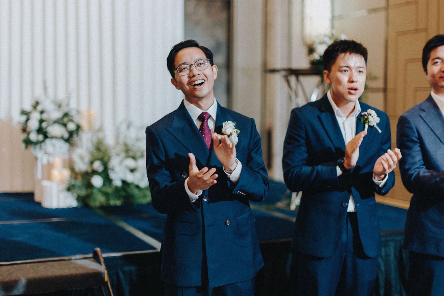 Wedding guests in navy suits applaud during an elegant indoor ceremony.