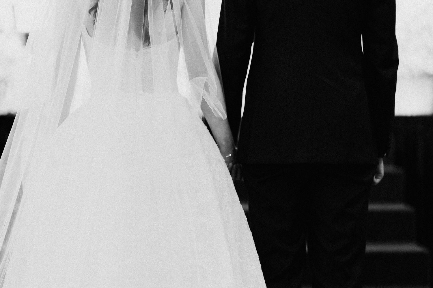 Black and white wedding photo of bride in white gown and veil holding hands with groom in black suit.