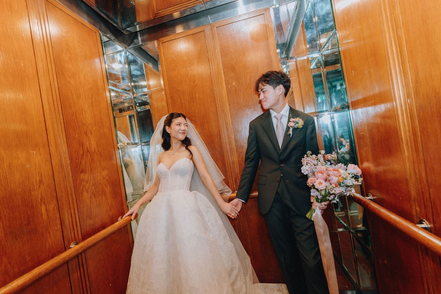 Newlywed couple holding hands and walking through elegant wood-paneled staircase with floral bouquet.