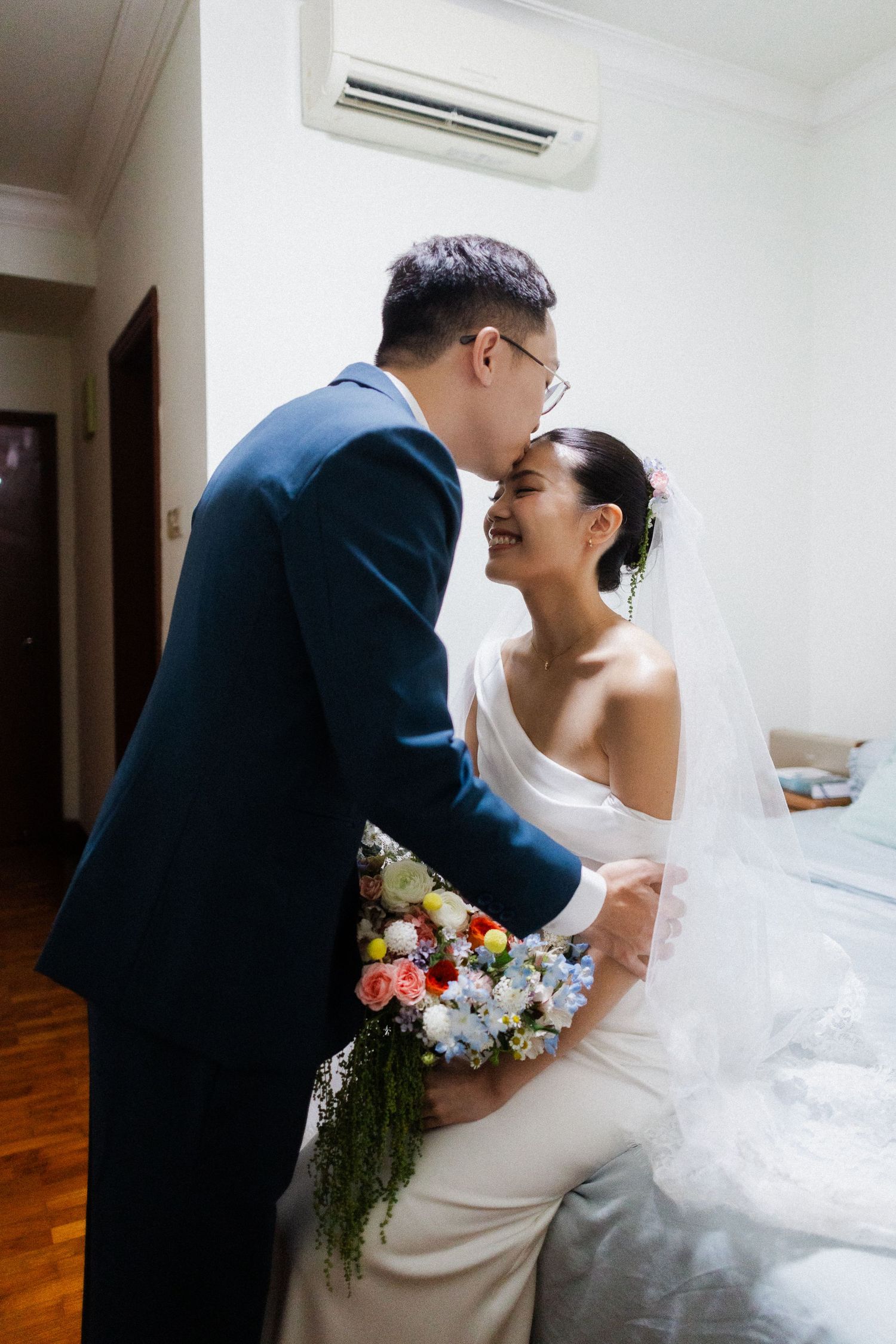Groom kissing bride on forehead as she holds colorful bouquet in a bright bedroom.