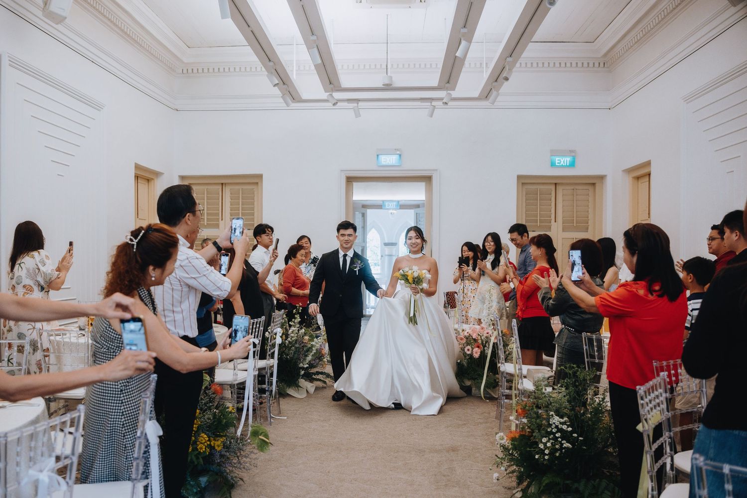 Wedding couple walks down aisle as guests celebrate and photograph the ceremony in a bright elegant venue.