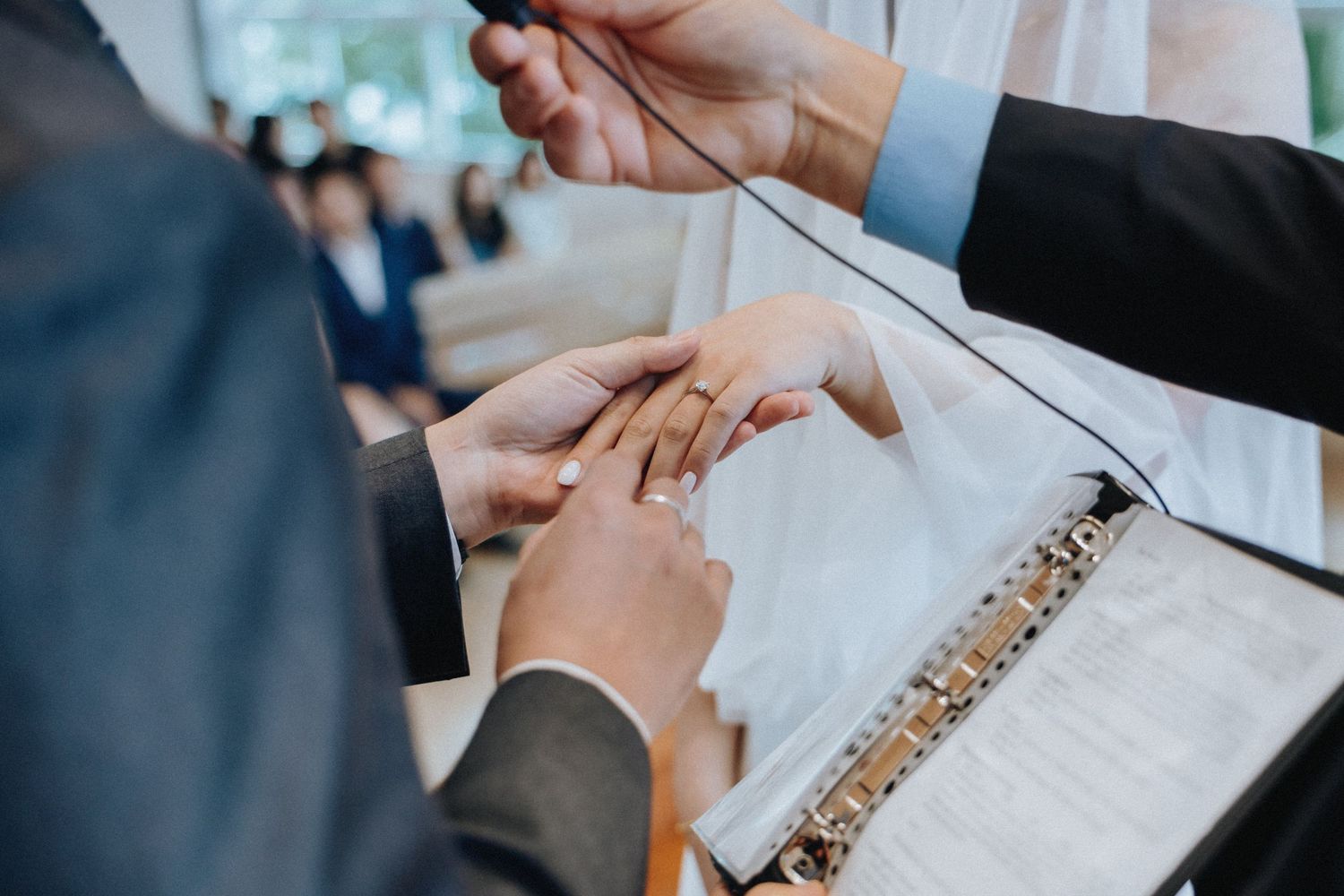 Close-up of wedding ring exchange ceremony with open Bible and officiant guiding couple hands.