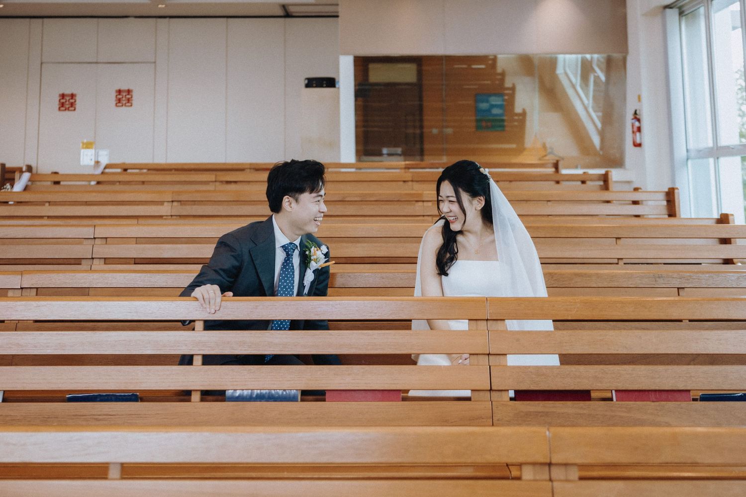 Bride and groom sit together in wooden church pews, smiling at each other before the ceremony.