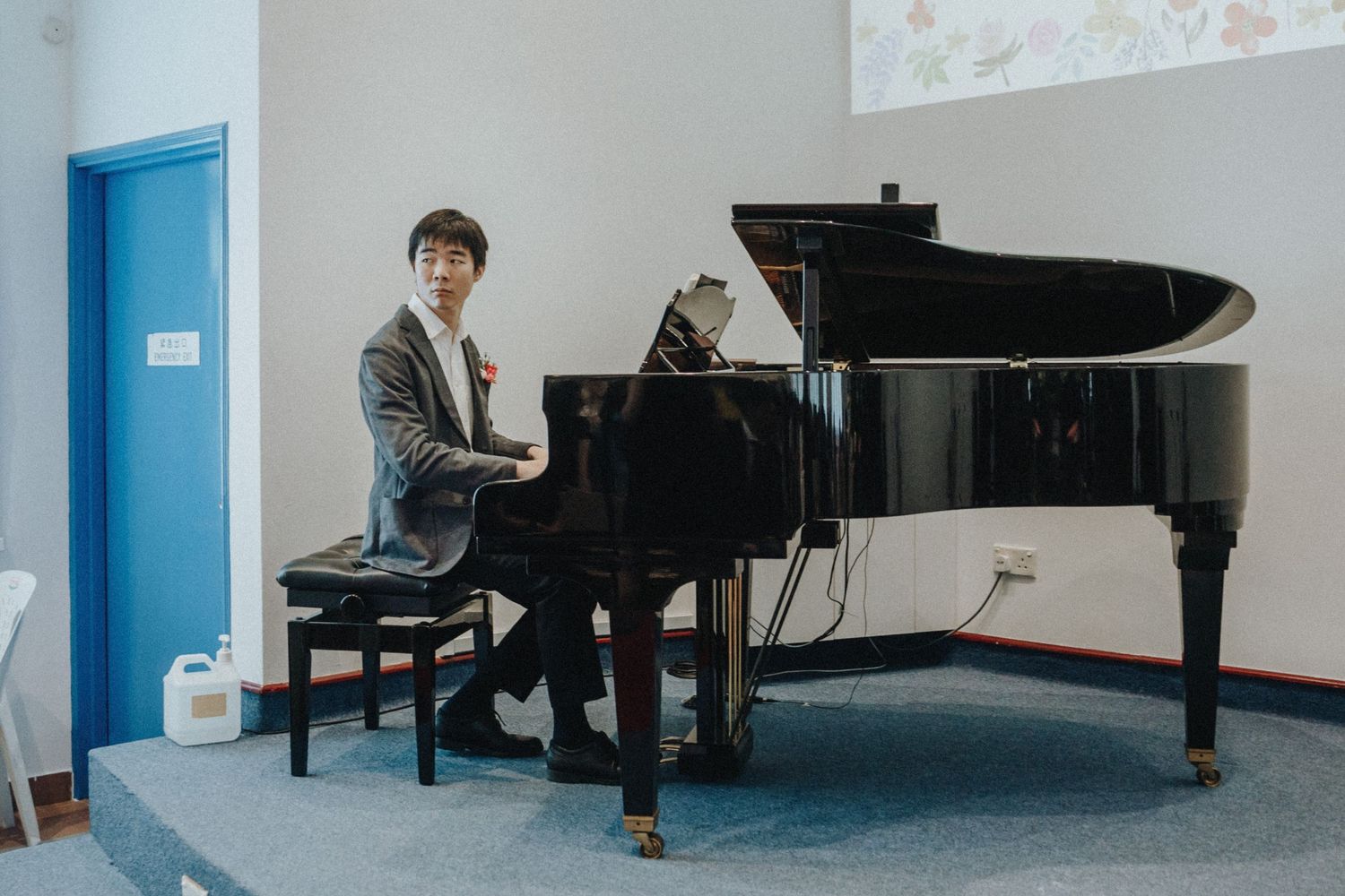 Young Asian man in gray blazer seated at a grand piano in a bright room with blue accents.
