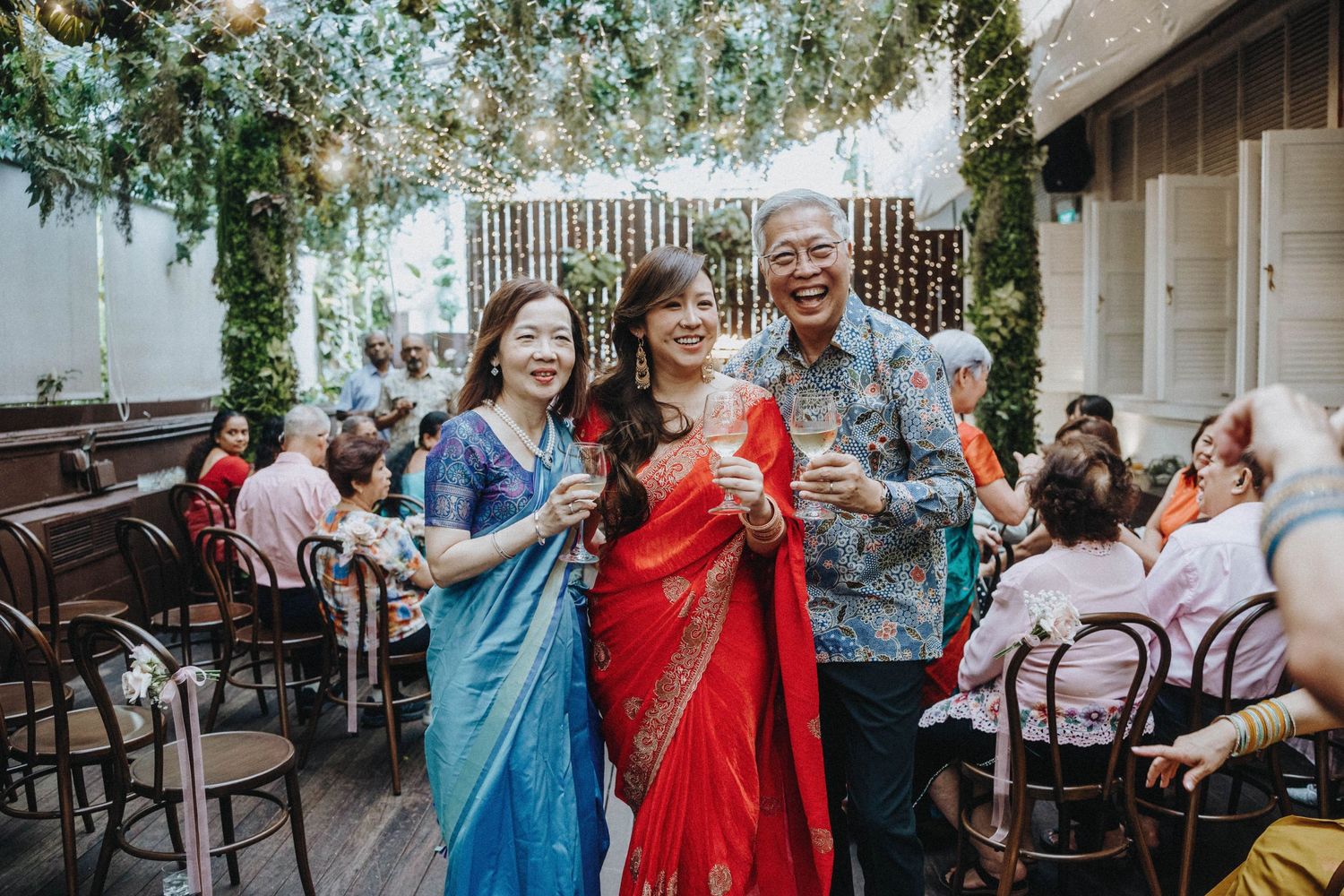Three smiling wedding guests in colorful Indian attire raising glasses in a toast at an outdoor reception.