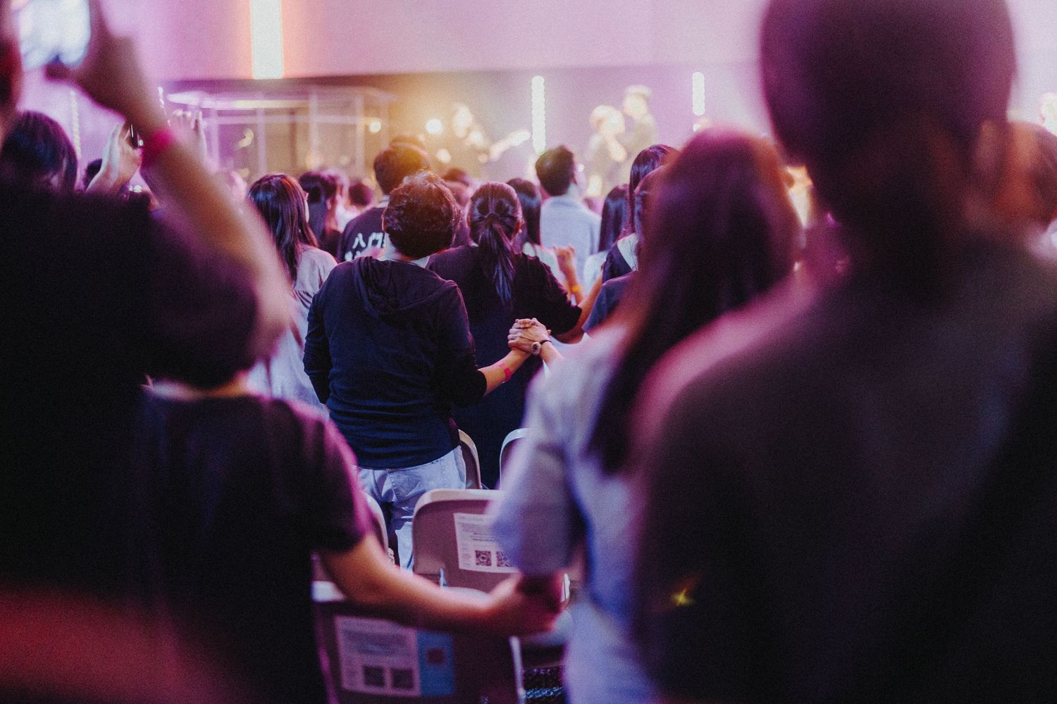 Crowd of concert attendees with raised hands in purple-tinted venue lighting.