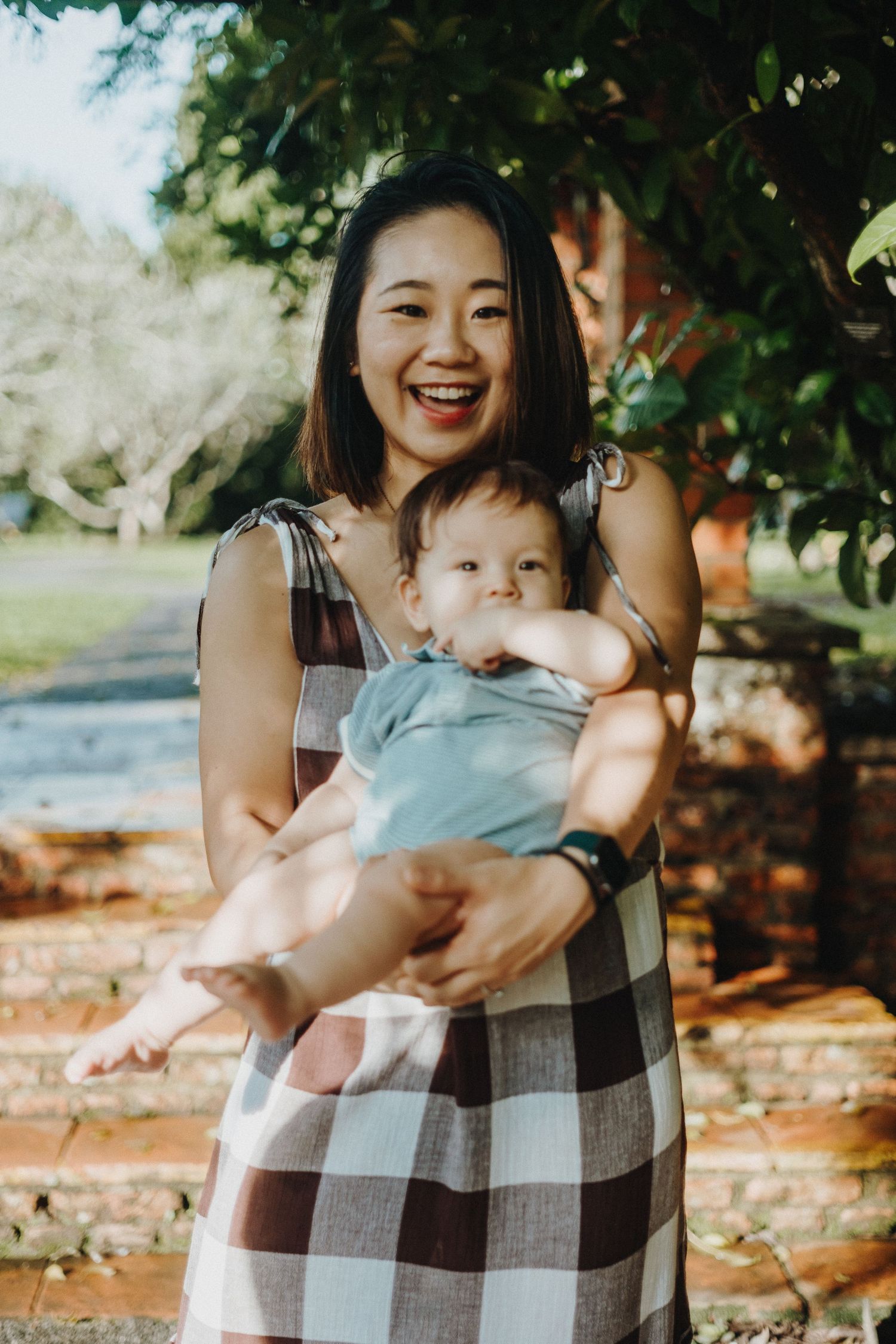 Woman in plaid dress holds baby outdoors against brick wall background in sunlit garden setting.