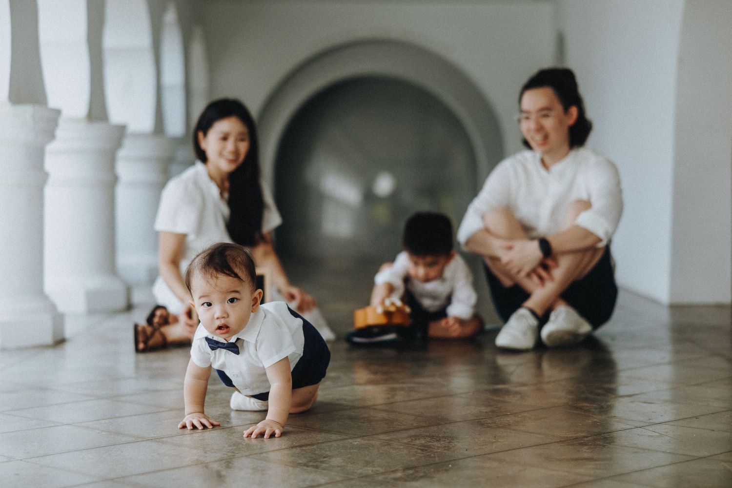 Family sitting together on a wooden floor in a hallway with white columns as a child crawls on the ground.