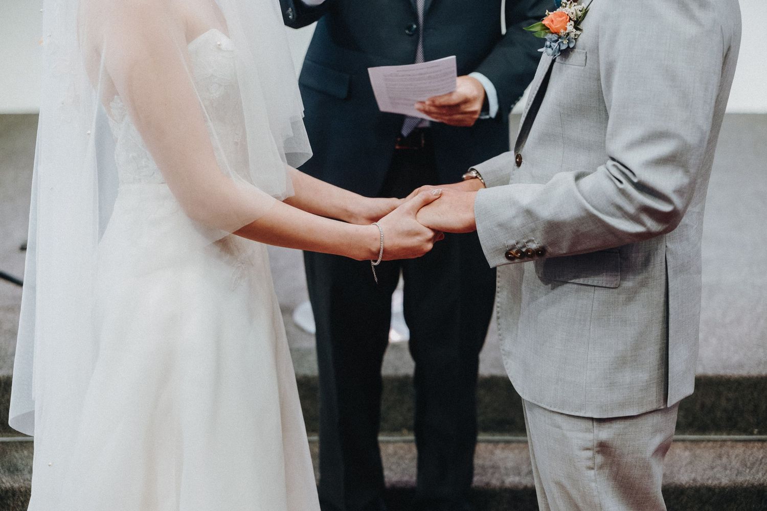 A wedding ceremony moment showing joined hands of the couple with an officiant in black holding papers.