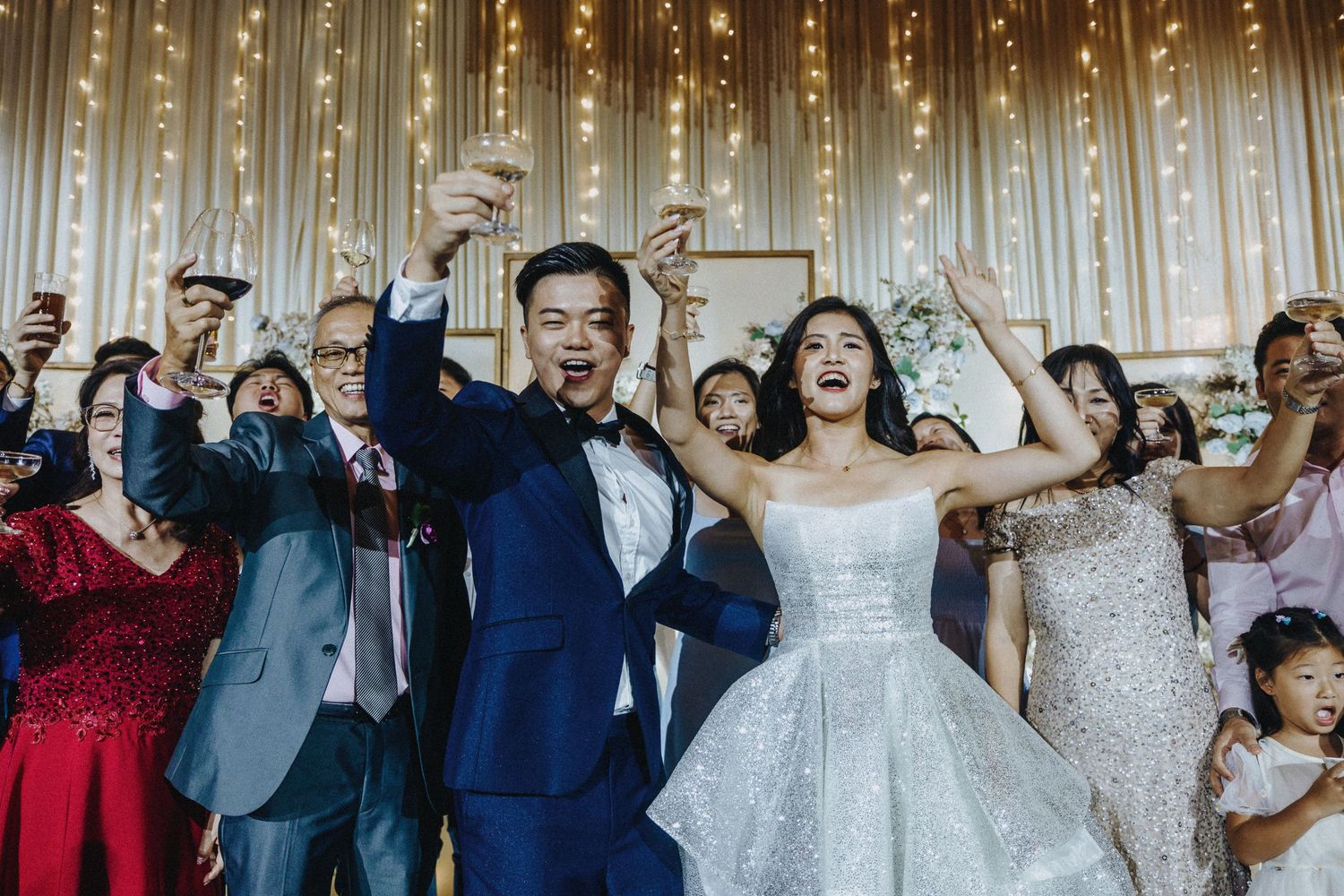 Joyful wedding celebration with guests cheering and raising hands against a sparkly backdrop.