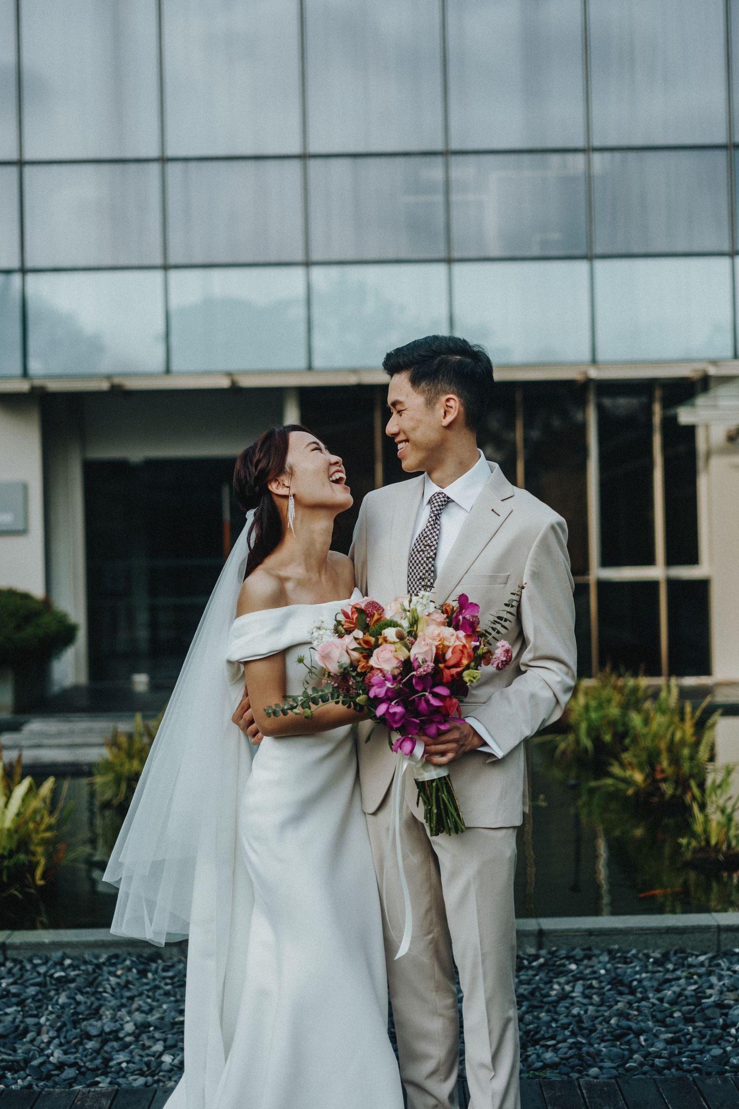 A bride and groom share a joyful moment outside a modern building with a colorful bouquet.