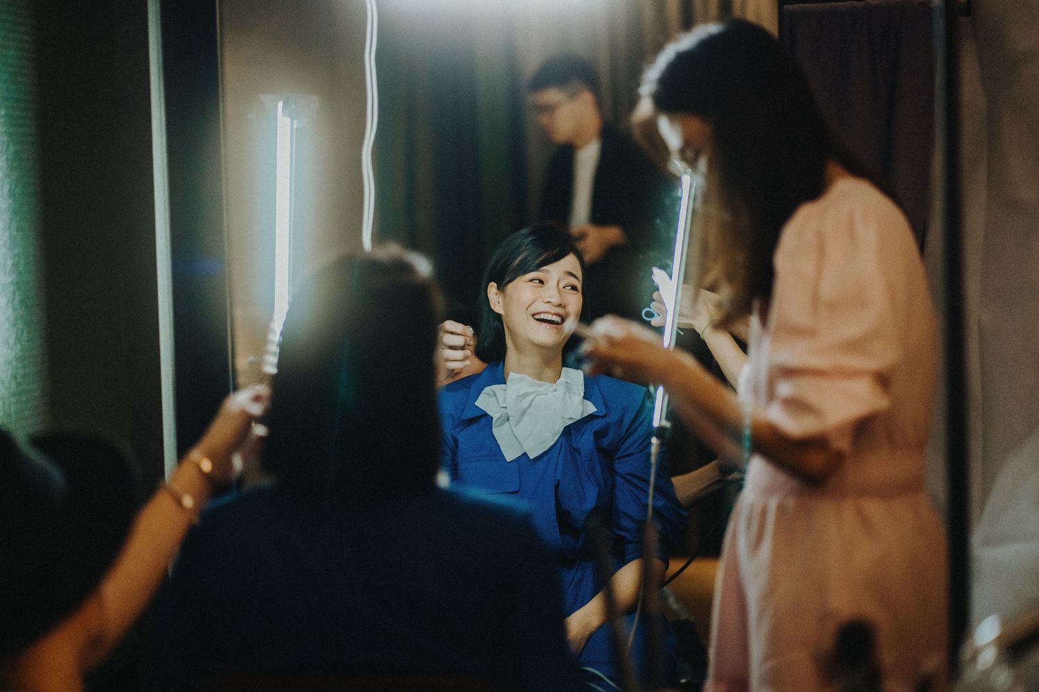 A group of people gathered backstage getting prepared with hair and makeup under bright lighting.