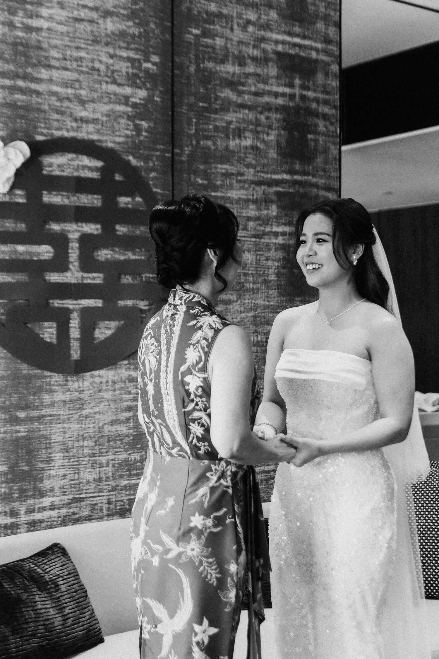 Women in festive patterned dresses stand together against a brick wall at a wedding celebration.