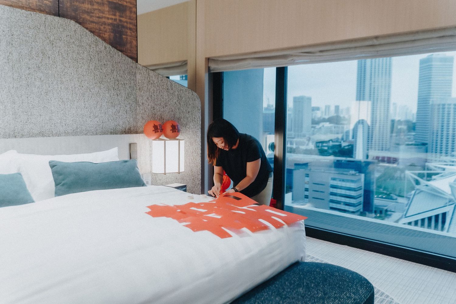 A housekeeper makes a bed in a modern hotel room with floor-to-ceiling windows overlooking a city skyline.