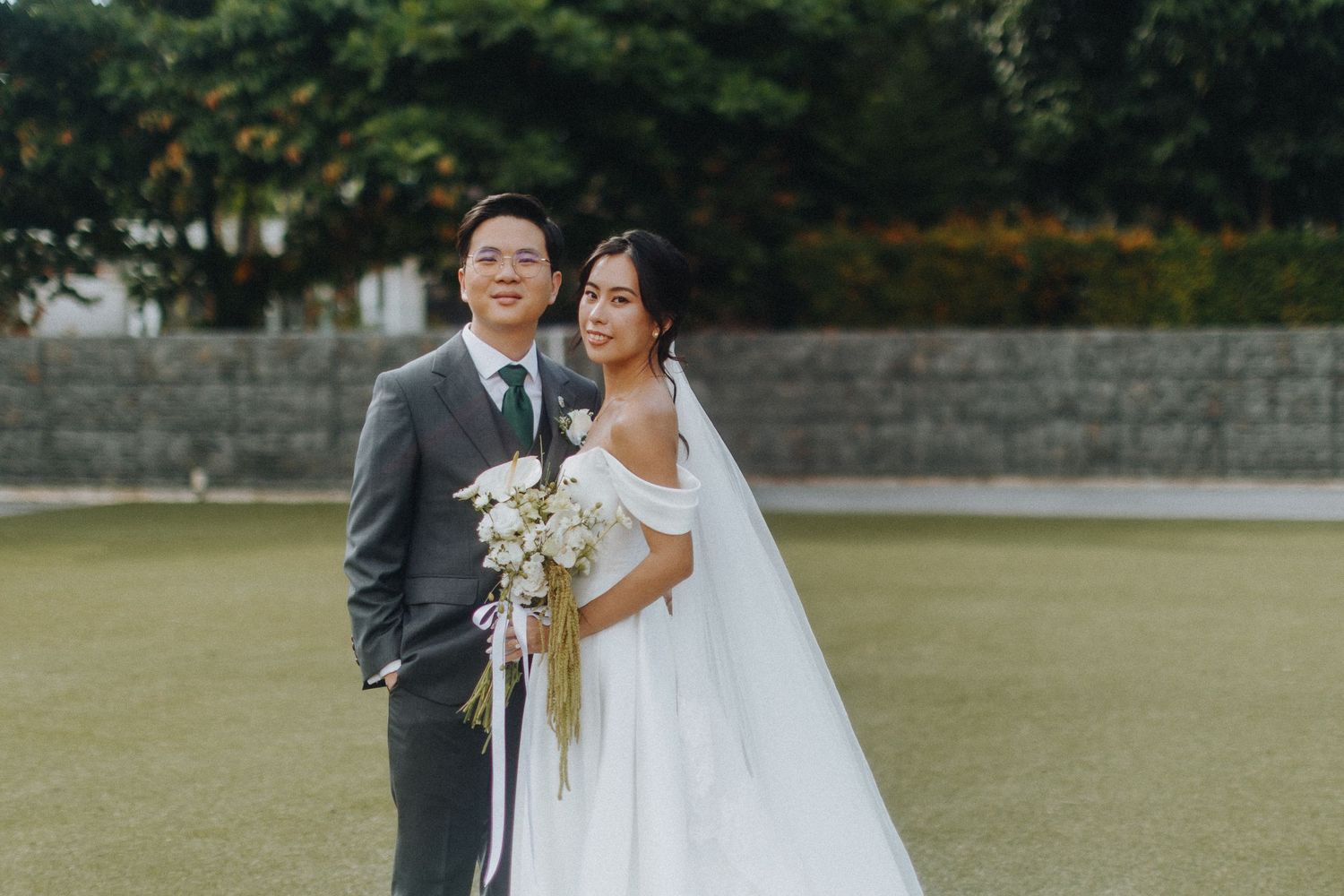 Wedding couple stands together on grassy lawn near stone wall during outdoor celebration.
