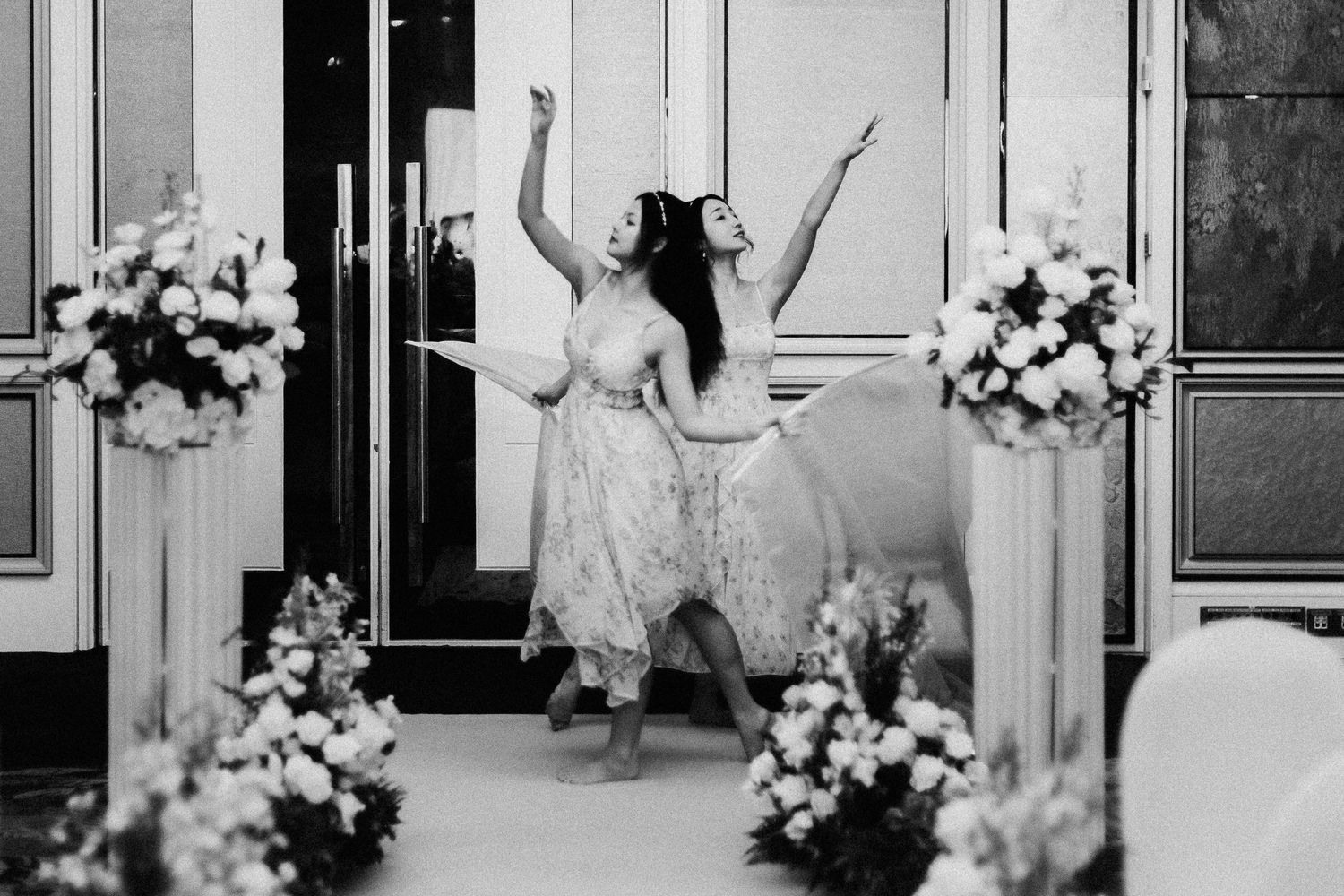 Two people dancing joyfully in white dresses surrounded by floral arrangements at an elegant venue.