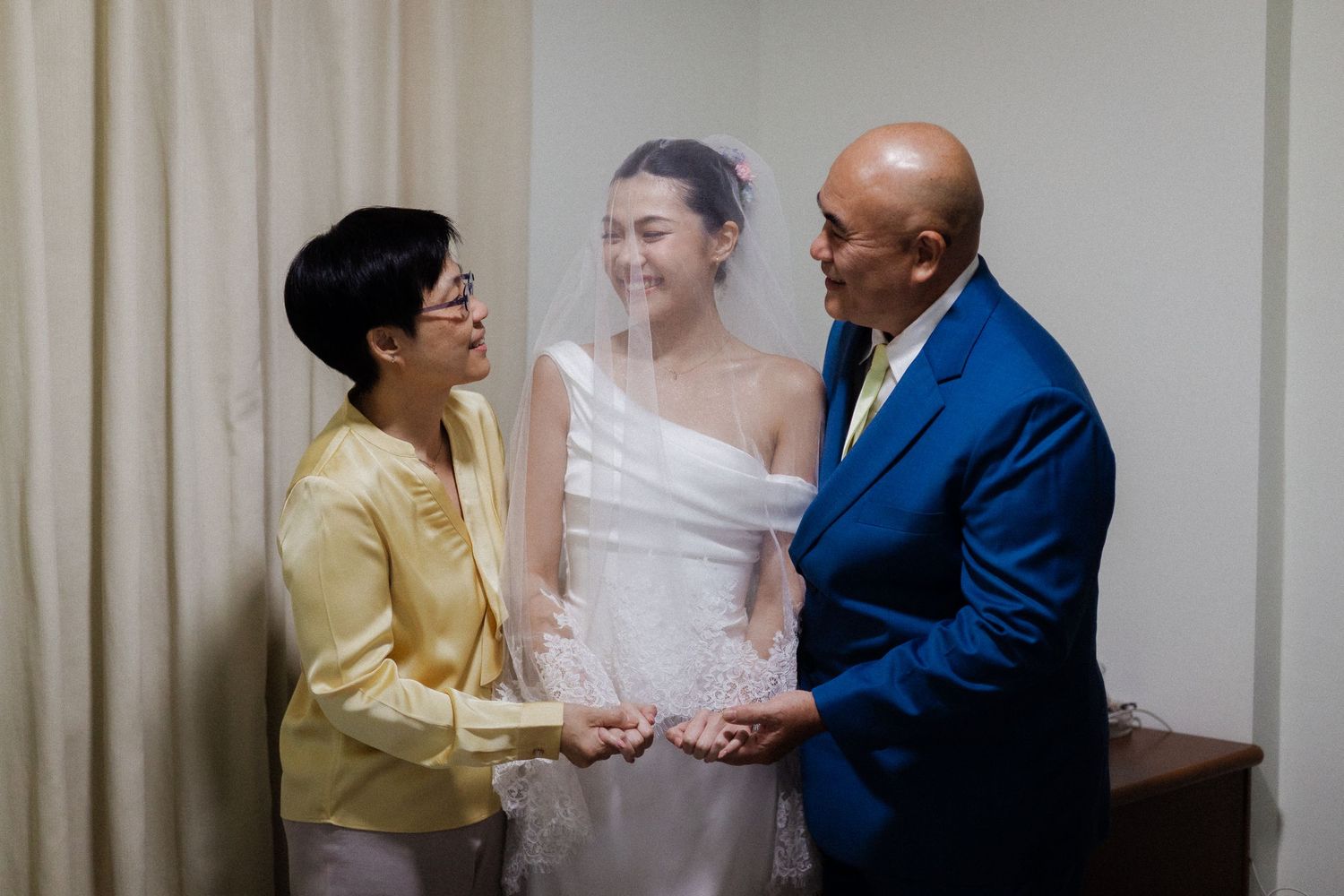Bride in white wedding dress with veil smiling between proud parents in formal attire indoors.