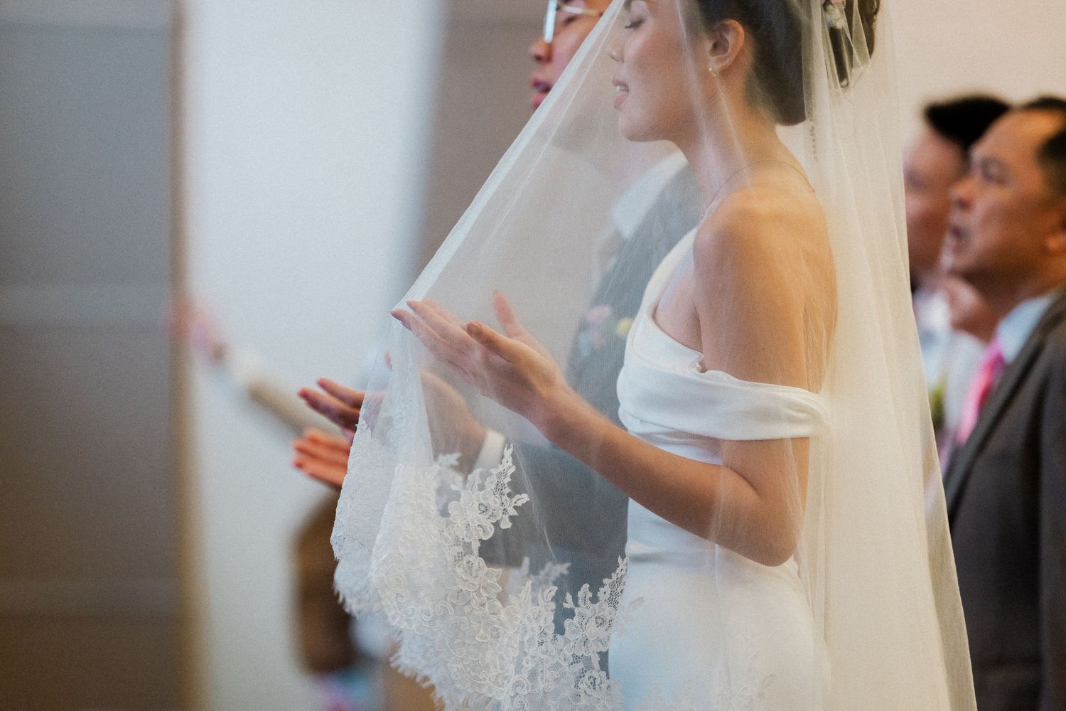 Bride in off-shoulder lace wedding gown and veil praying during wedding ceremony.