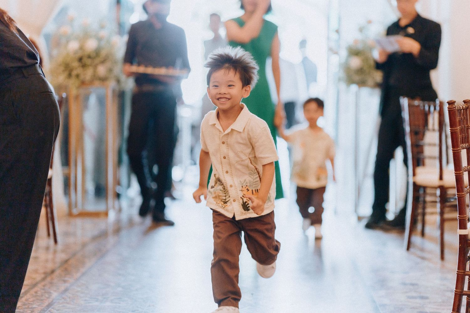 A joyful young boy in a cream shirt and brown pants runs down a wedding aisle, smiling brightly at an elegant venue.