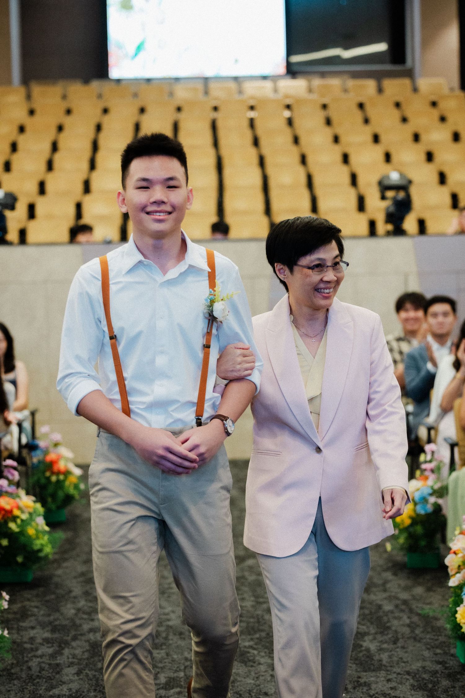 Smiling groom in suspenders walks down wedding aisle escorted by mother in pink blazer.